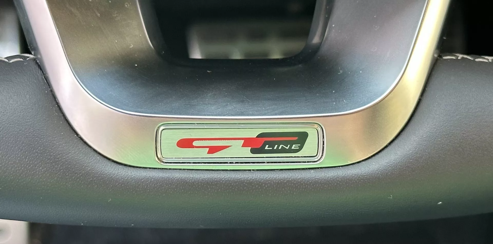 Posnetek iz bližine emblema volana Kia XCeed 2022 GT-Line, ki prikazuje kovinske in črne črke z športnim rdečim poudarkom, obdan z vrhunskim usnjenim in kovinskim volanom
