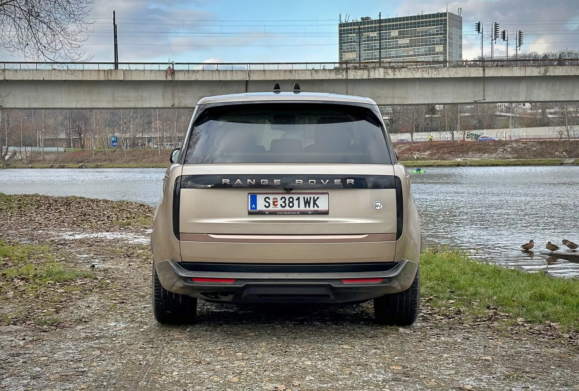 Zadnji pogled na Range Rover 2021 z neposrednim kotom pogleda na ravni oči, parkiran v bližini vodnega telesa z račkami in nadvozom v ozadju, prikazuje ime modela, zadnje luči, senzorje za parkiranje in dvojni izpušni sistem v mat bež barvi