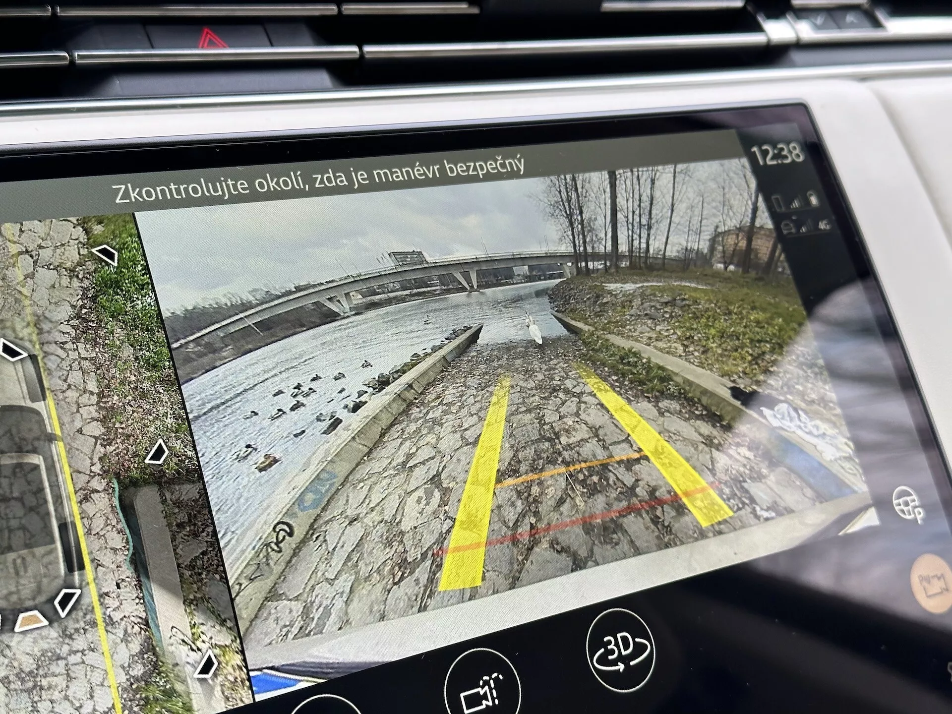 Zaslon informacijsko-zabavnega sistema Land Rover Range Rover 2021, ki prikazuje zadnji pogled iz kamere za vzvratno vožnjo z dinamičnimi vodilnimi črtami na parkirišču blizu vodne površine, podrobno z vegetacijo, ikonami pomoči pri parkiranju in besedilom varnostnega opomnika