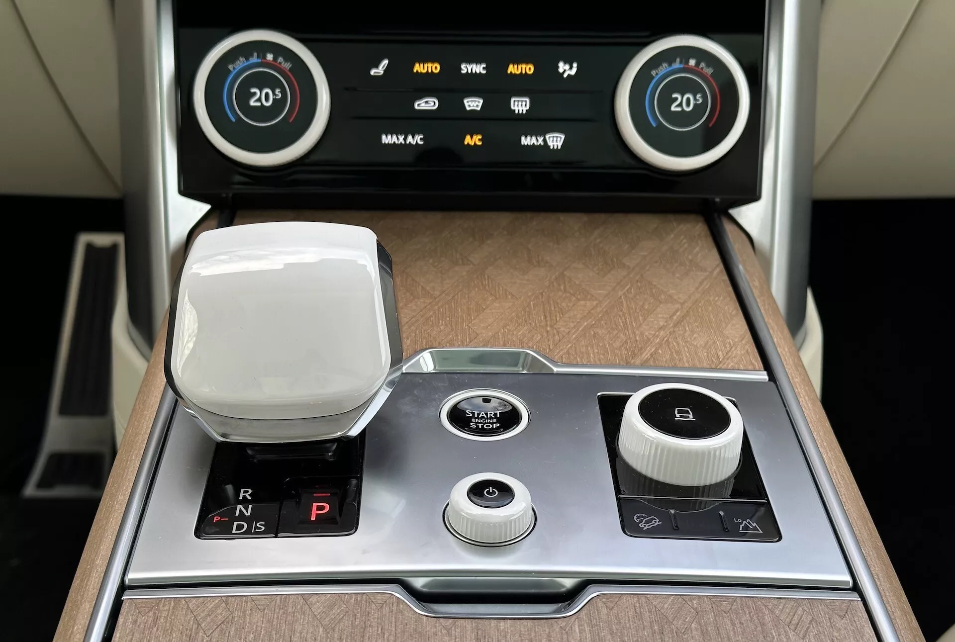 Podroben pogled na sredinsko konzolo Land Rover Range Rover 2021 z elektronskim izbirnikom prestav, ki prikazuje oznake za vzvratno, nevtralno, parkirno in vožnjo, gumb za elektronsko ročno zavoro, večnamenski vrtljivi gumb za sistem zabave, gumb za nadzor glasnosti, gumb za zagon/zaustavitev motorja, in zaslon za dvoconsko avtomatsko klimatsko napravo z lesnimi in kovinskimi okraski