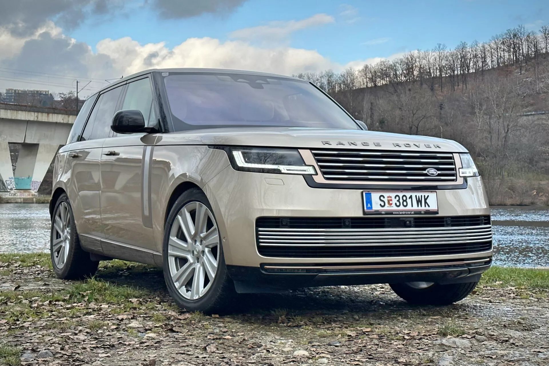 Luksuzni SUV Land Rover Range Rover 2021, pogled s sprednje in treh četrt strani, ki prikazuje kromirano masko, LED žaromete, večkraka lita kolesa in aerodinamično zasnovo, parkiran na prostem blizu reke in mostu