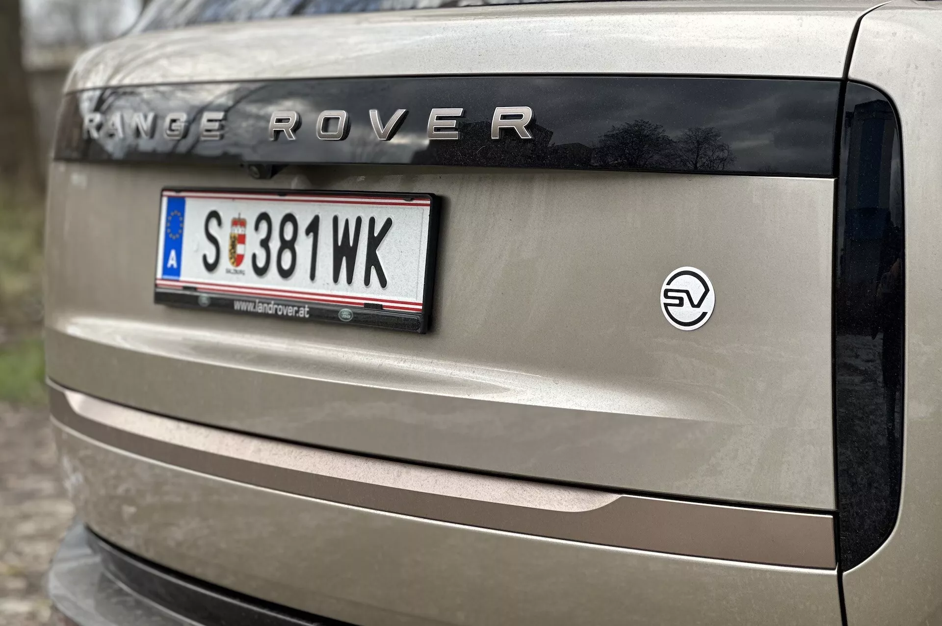 Podroben pogled na zadnji del vozila Range Rover 2021 z oznako 'RANGE ROVER', evropskim nosilcem registrske tablice, emblemo EV in delom zadnje luči