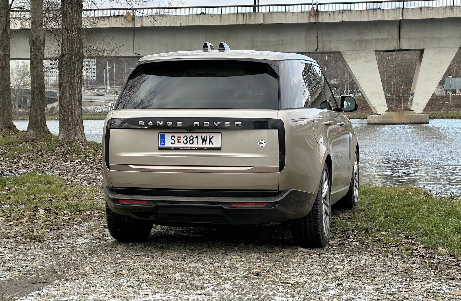 Zadnji pogled na Range Rover 2021, parkiranega na prostem z vodnim telesom in mostom v ozadju, prikazuje LED zadnje luči, dvojni izpuh, stekla za zasebnost in večkraka lita platišča.