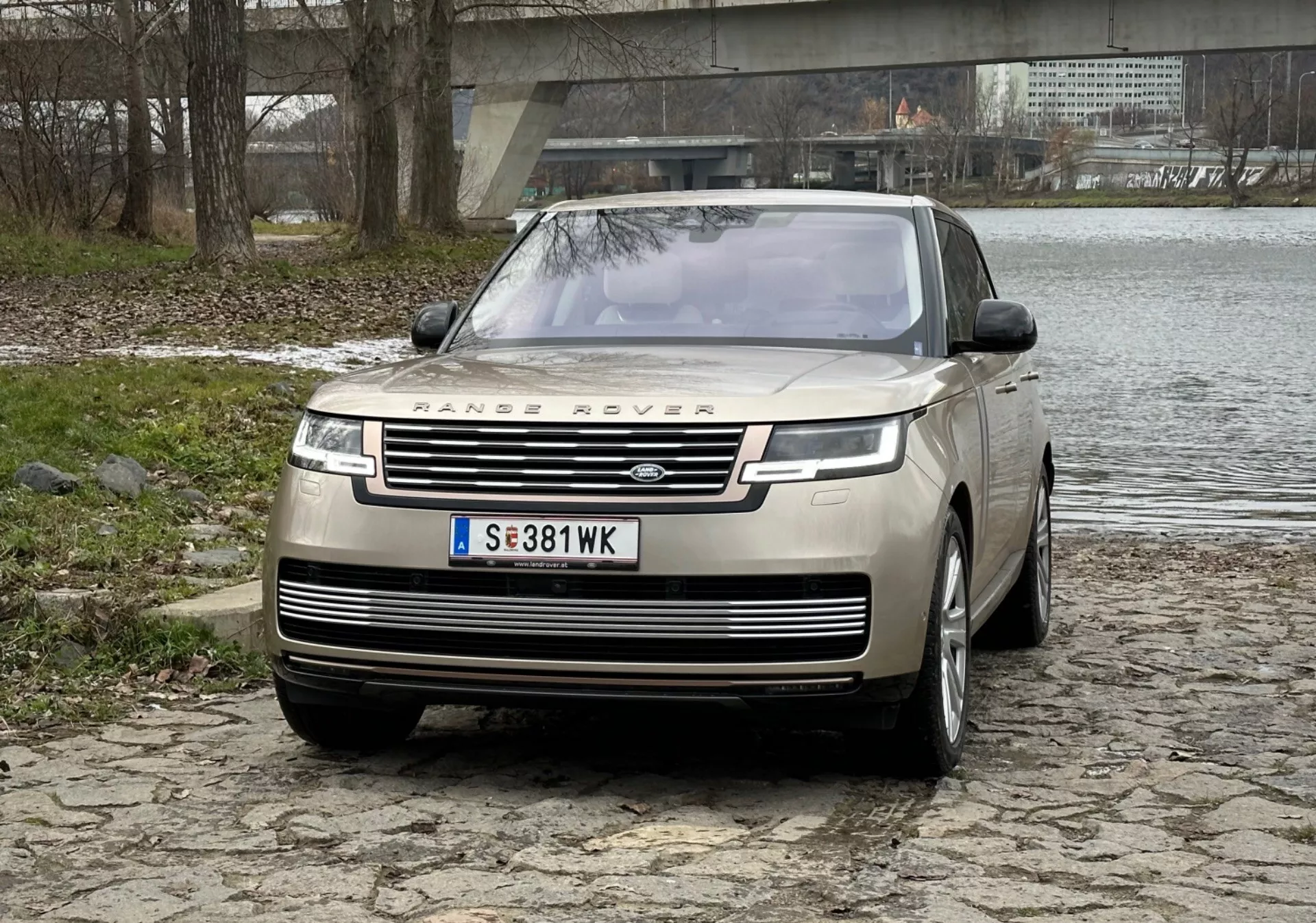 Luksuzni SUV Range Rover 2021 v srebrni barvi, poudarja izstopajočo sprednjo masko z blagovno znamko, elegantne LED dnevne luči, sprednji odbijač s parkirnimi senzorji in dizajn motornega pokrova clamshell na tlakovani površini ob reki