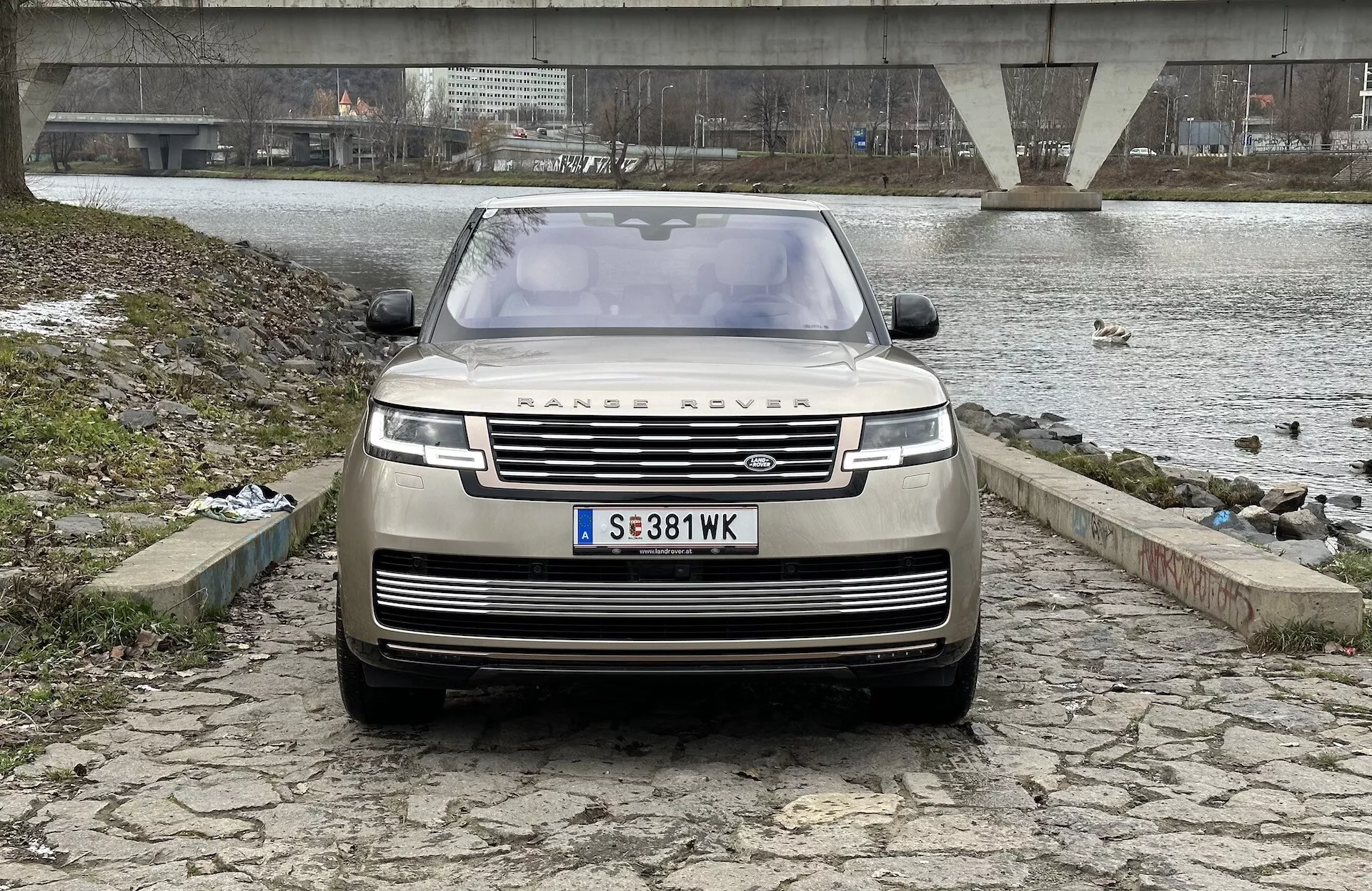 Land Rover Range Rover 2021, fotografiran iz sprednjega, nekoliko nizkega kota, ki poudarja rešetko, sprednji odbijač in pokrov motorja, z ikoničnim logotipom RANGE ROVER, sodobnimi LED žarometi in napredno tehnologijo pomoči vozniku, s pogledom na reko ali jezero, most in oblačno nebo