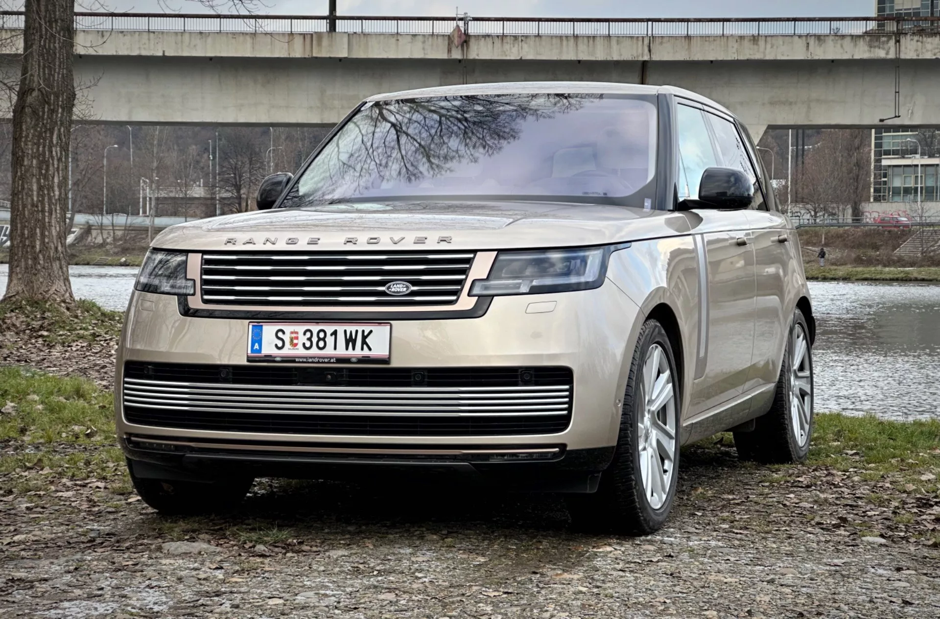 Range Rover 2021 v impresivnem pogledu s sprednje tričetrtinske strani, ki prikazuje njegovo značilno rešetko, elegantne LED žaromete in robustno sprednjo zaščitno ploščo proti ozadju mestnega parka, poudarjajoč luksuznost in zmogljivost za terensko vožnjo.