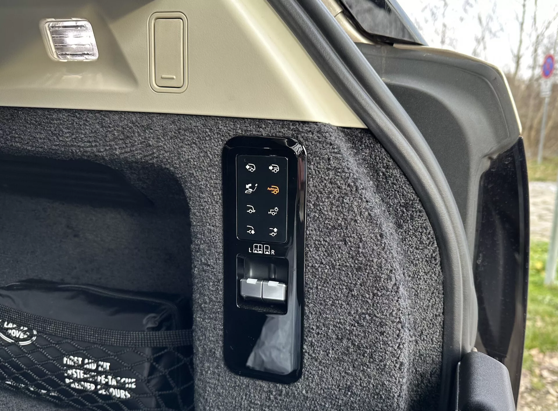 Pogled od blizu na elektronsko nadzorno ploščo prtljažnika v notranjosti Land Rover Range Roverja 2021, ki prikazuje gumbe za operacije kot so elektronsko odpiranje in zapiranje, nadzor zložljivega prtljažnega pokrova, možno svetlo prtljažnika ali pomožno vtičnico, nastavitve zadnjih sedežev iz prtljažnika in izredno sprostitev, nastavljeno proti preprogi bočne stene prtljažnika in svetlemu stropu s svetlobnim telesom tovora.