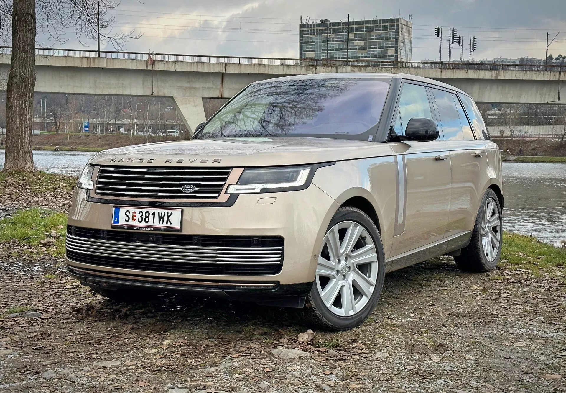 Luksuzni SUV Land Rover Range Rover 2021, parkiran na mešanem terenu zemlje in trave z betonsko konstrukcijo in vegetacijo v ozadju, prikazuje pogled sprednjega kota, ki poudarja LED žaromete, lita kolesa in moderne aerodinamične lastnosti