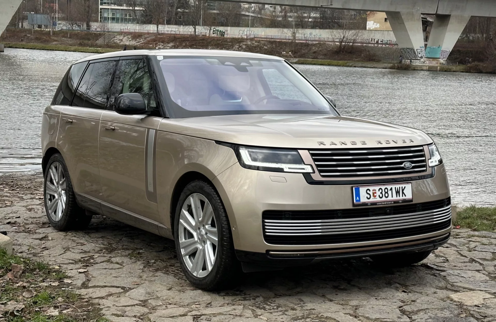 Luksuzni SUV Land Rover Range Rover letnika 2021 parkiran ob rečni obali z mostom v ozadju, ki prikazuje svoj sodoben dizajn in tehnološke značilnosti, vključno z LED žarometi, sprednjo rešetko z emblemo, večkraka lita kolesa in panoramsko strešno okno.