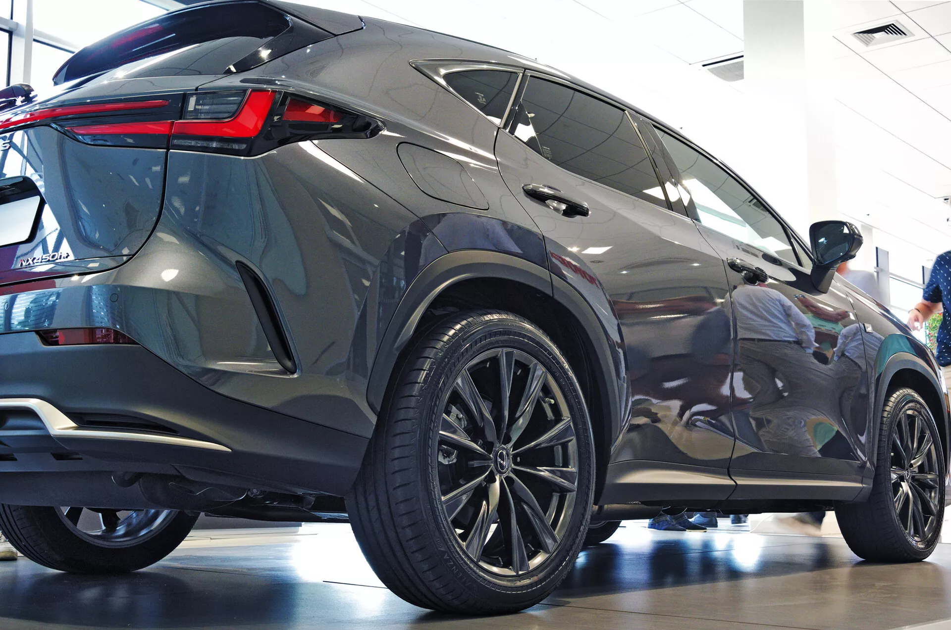 Zadnji tričetrtinski kot SUV Lexus NX 2021, ki prikazuje bočni profil in zadnji del z velikimi večkrakimi litimi platišči, nizkoprofilnimi pnevmatikami, ostrimi zadnjimi LED lučmi, kovinsko karoserijo, zatemnjenimi okni, integriranih zadnjim spojlerjem ter čistem in sijočim salonskim okoljem, ki poudarja moderno estetiko in luksuzne lastnosti