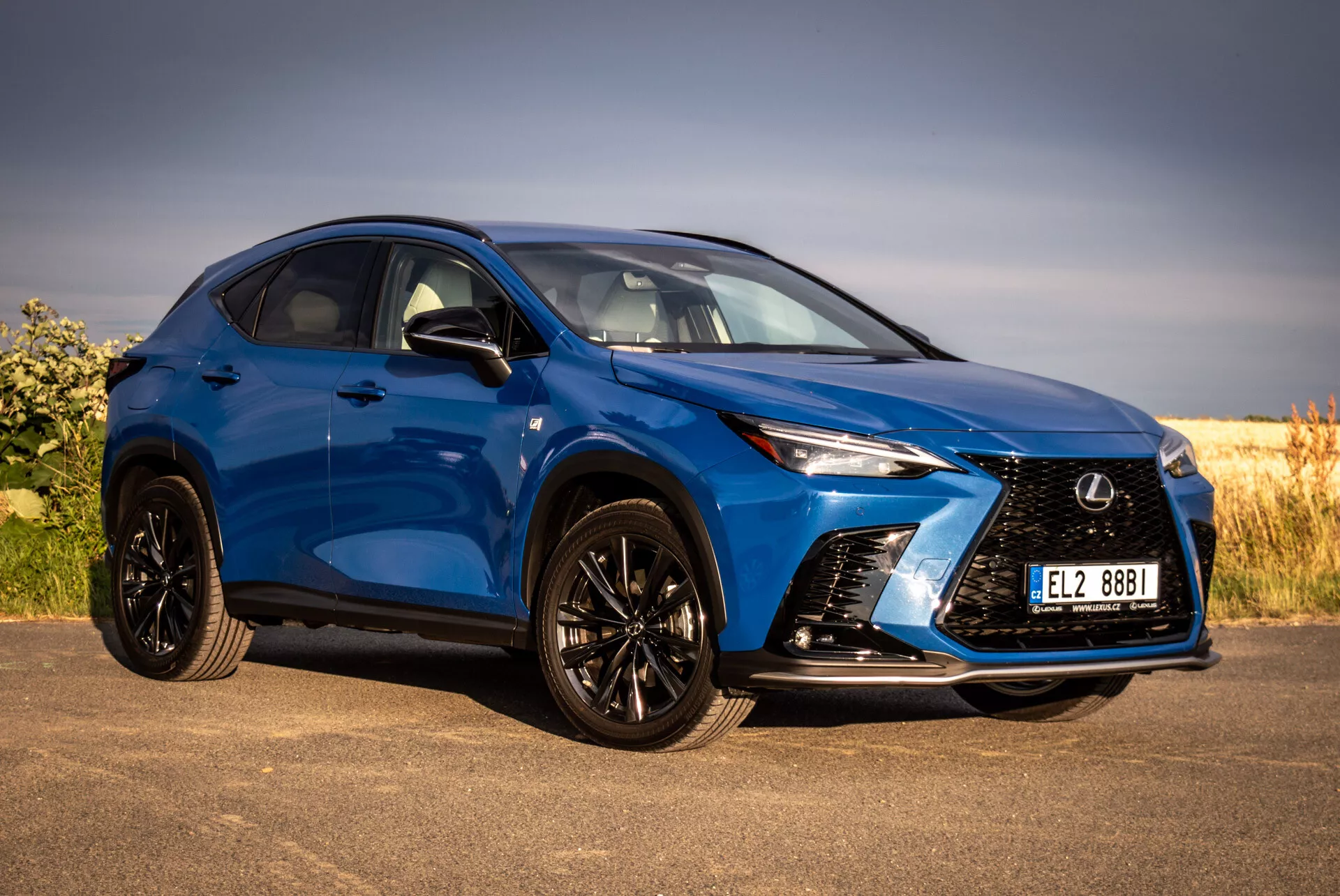 Modri SUV Lexus NX 2021 v pogledu s sprednje strani tri četrtine, ki poudarja njegov agresiven žaromet, ostre LED žaromete, črna lita kolesa z nizkoprofilnimi pnevmatikami in ogledala ob strani v barvi karoserije, proti naravnemu ozadju