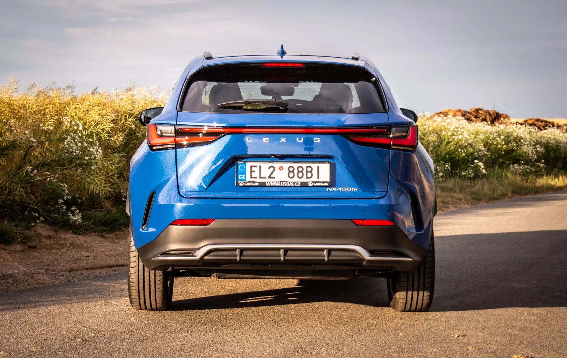 Zadnji pogled na modri Lexus NX 2021 iz nizkega kota, ki poudarja trdno zgradbo in oblikovalske značilnosti avtomobila, kot so LED zadnje luči, zadnje vetrobransko steklo, spojler, očitno značenje, zadnji odbijač z difuzorjem, parkirni senzorji, odsevniki in dvojni izpuh, na naravnem ozadju pod jasnim nebom.
