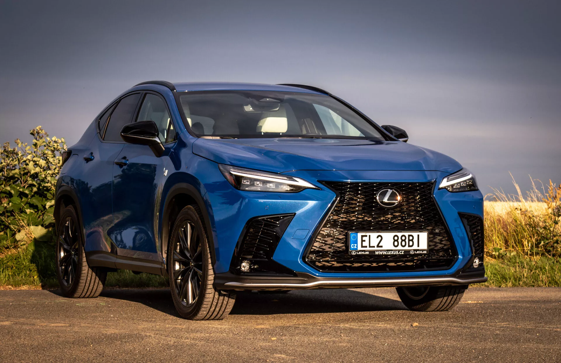 Posnetek iz nizkega kota modrega SUV-ja Lexus NX 2021, ki prikazuje sprednjo rešetko z dizajnom vretena, LED žaromete, črna lita kolesa in integrirane smernike na stranskem ogledalu, ki poudarjajo njegove luksuzne in zmogljive lastnosti.