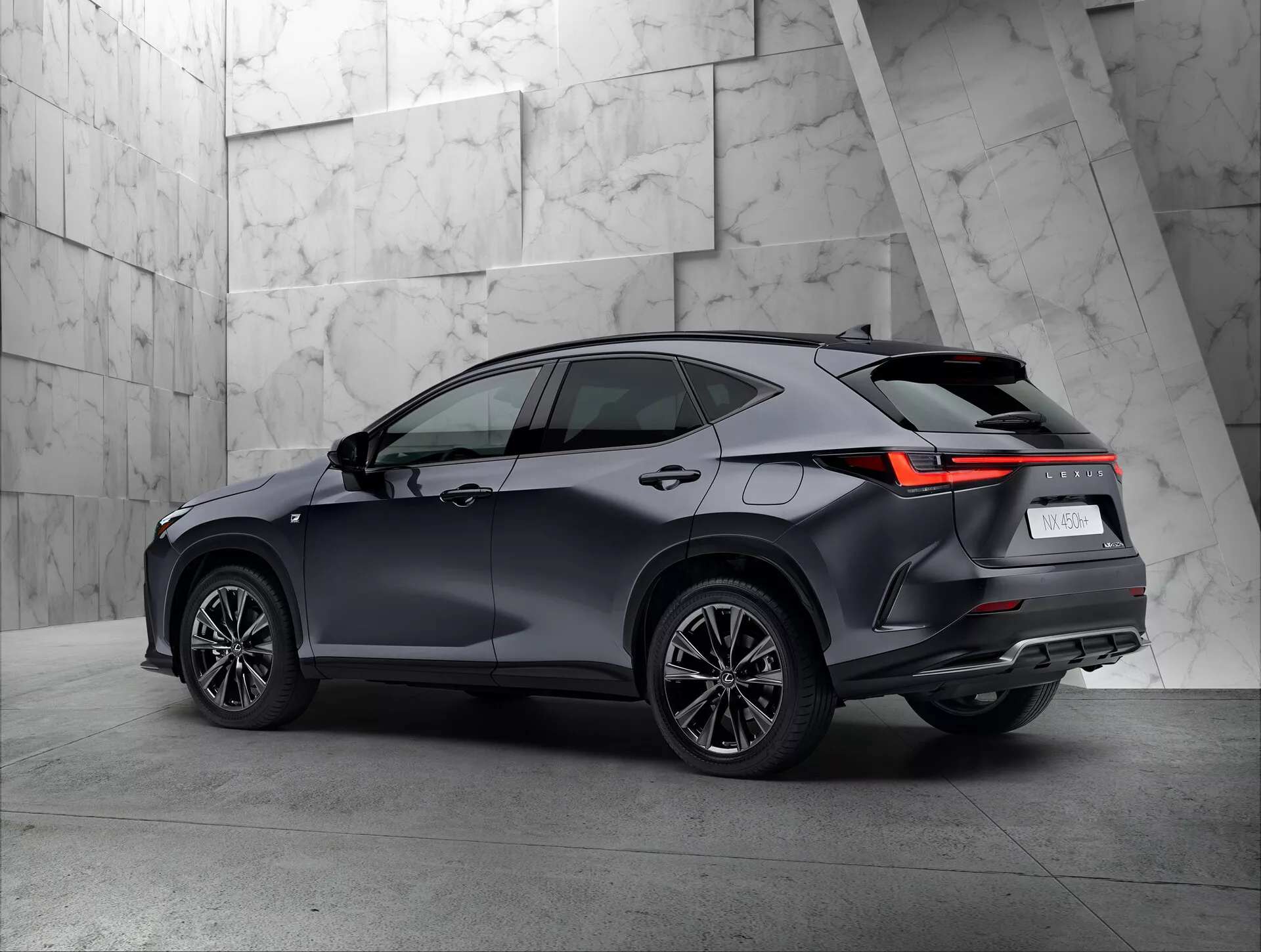 Tričetrtinski zadnji pogled na sodoben SUV Lexus NX 2021 v razstavnem salonu. Vozilo jasno prikazuje desno stran in zadnji del, vključno z zadnjimi lučmi z LED dizajnom, temno sijočo barvo, večkrakimi kolesi in strešnim linijo z možnimi strešnimi nosilci. Nevtralno, teksturirano ozadje lepo kontrastira s sofisticiranim dizajnom SUV.