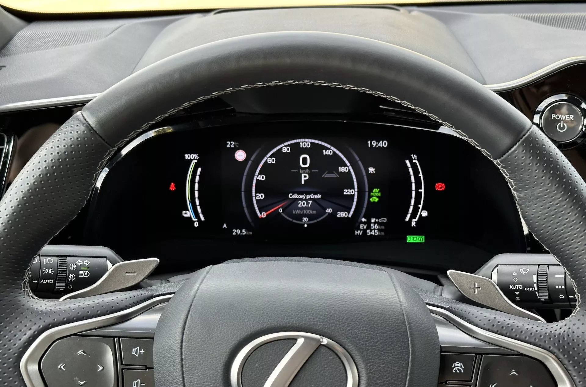 Pogled iz voznikovega sedeža na volan in digitalno armaturno ploščo Lexus NX 2021, poudarjanje indikatorjev hibridne tehnologije, gumbov za nadzor udobja in ergonomskega oblikovanja notranjosti
