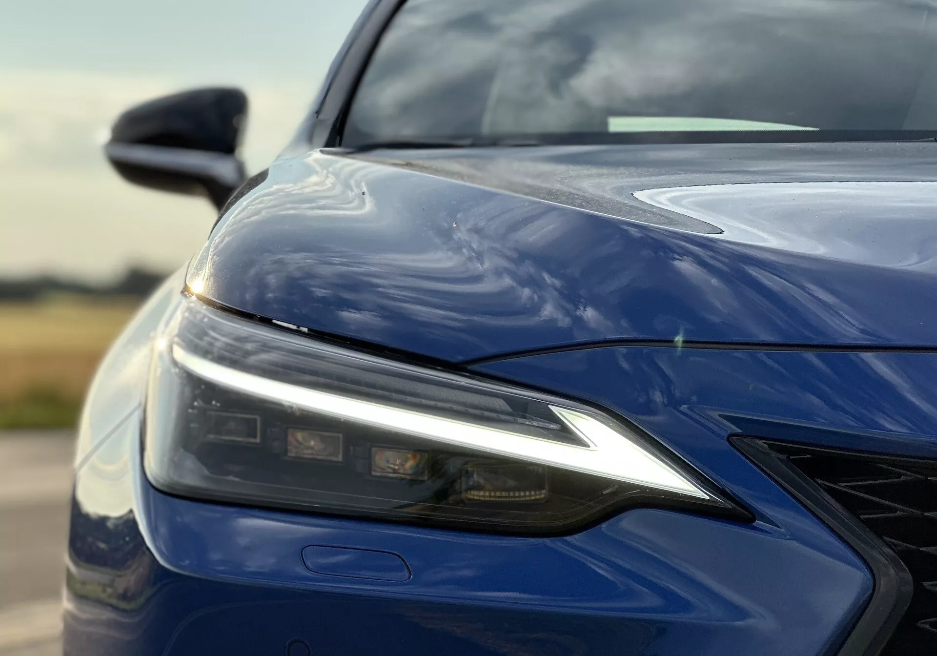 Od blizu prikazano levo sprednje četrtino Lexusa NX 2021 z LED žarometom, oblikovanim pokrovom motorja, sprednjim odbijačem z rešetko in sijajno globoko modro barvo, poudarja napredno tehnologijo razsvetljave in aerodinamično zasnovo.