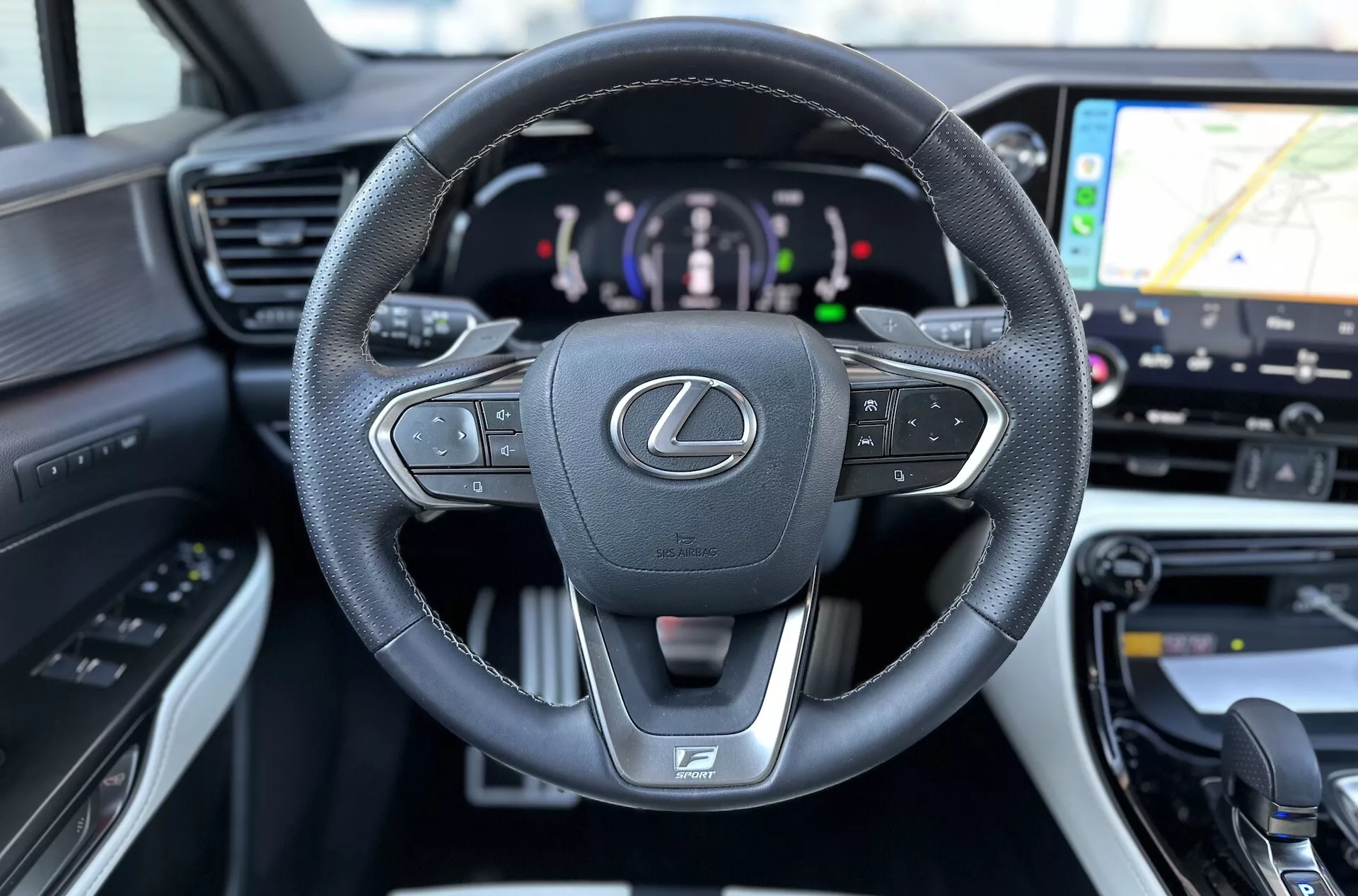 Notranjost Lexusa NX 2021 s poudarkom na usnjenem, multifunkcionalnem volanu z emblemo Lexus in logotipom F SPORT, digitalni instrumentalni plošči, ki kaže na hibridni pogon, in pogled na GPS navigacijo na sistemu za informacije in zabavo