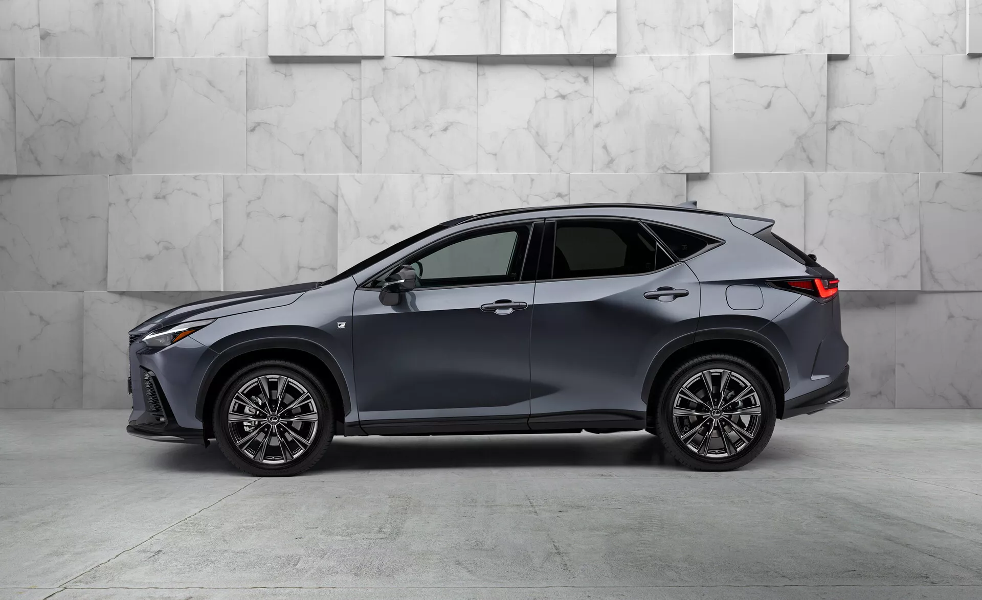 Stranski profil sodobnega crossover SUV vozila Lexus NX 2021, ki prikazuje povečano oddaljenost od tal in obliko karoserije. Vozilo je pobarvano v temni barvi, verjetno sivi ali modri, ima pet vrat, eleganten aerodinamičen dizajn, velika večkraka lita platišča, tanke LED žaromete in zadnje luči, stranska ogledala v barvi karoserije z vgrajenimi smerniki, okna s kromiranimi obrobami in anteno v obliki plavuti morskega psa, vse skupaj na ozadju iz marmorja.