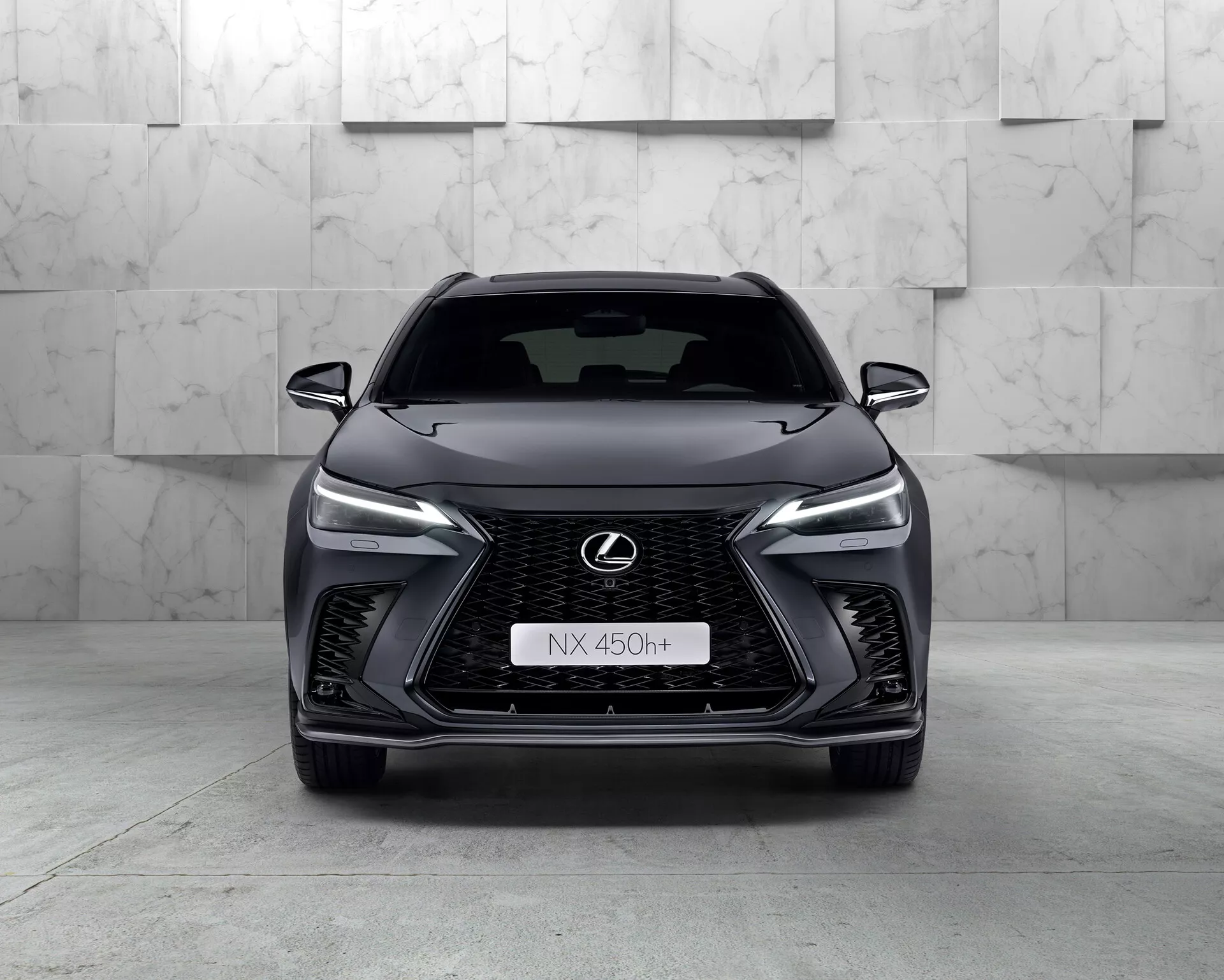 Čelni pogled na Lexus NX 450h+ 2021, eleganten sodoben SUV, ki poudarja svojo čelno masko v obliki vretena z vzorcem mreže, ozke LED žaromete, kromiran znak Lexus z modrim obrisom ter agresiven dizajn sprednjega odbijača. Hibridni značaj avtomobila je označen z znakom 'h+' na maski. Studijsko okolje poudarja obline in značilnosti vozila s čisto, kontrolirano osvetlitvijo.
