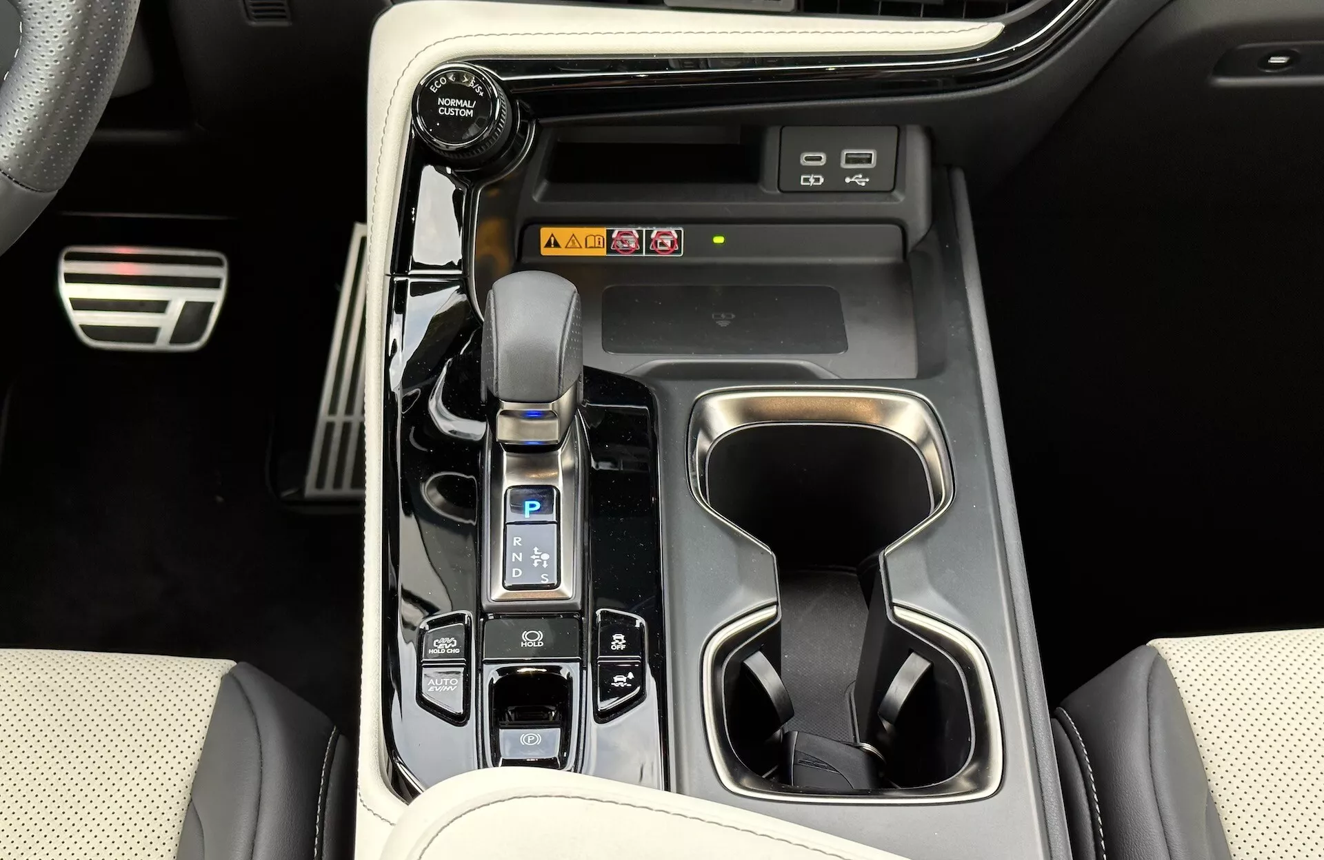 Pogled na sredinsko konzolo Lexus NX 2021, ki prikazuje samodejni menjalnik z usnjenim pokrovom, izbirnik načina vožnje, držala za kozarce, gumbe za nadzor vozila, možne bližnjice do sistema za zabavo in informacije ter elemente razkošnega notranjega oblikovanja