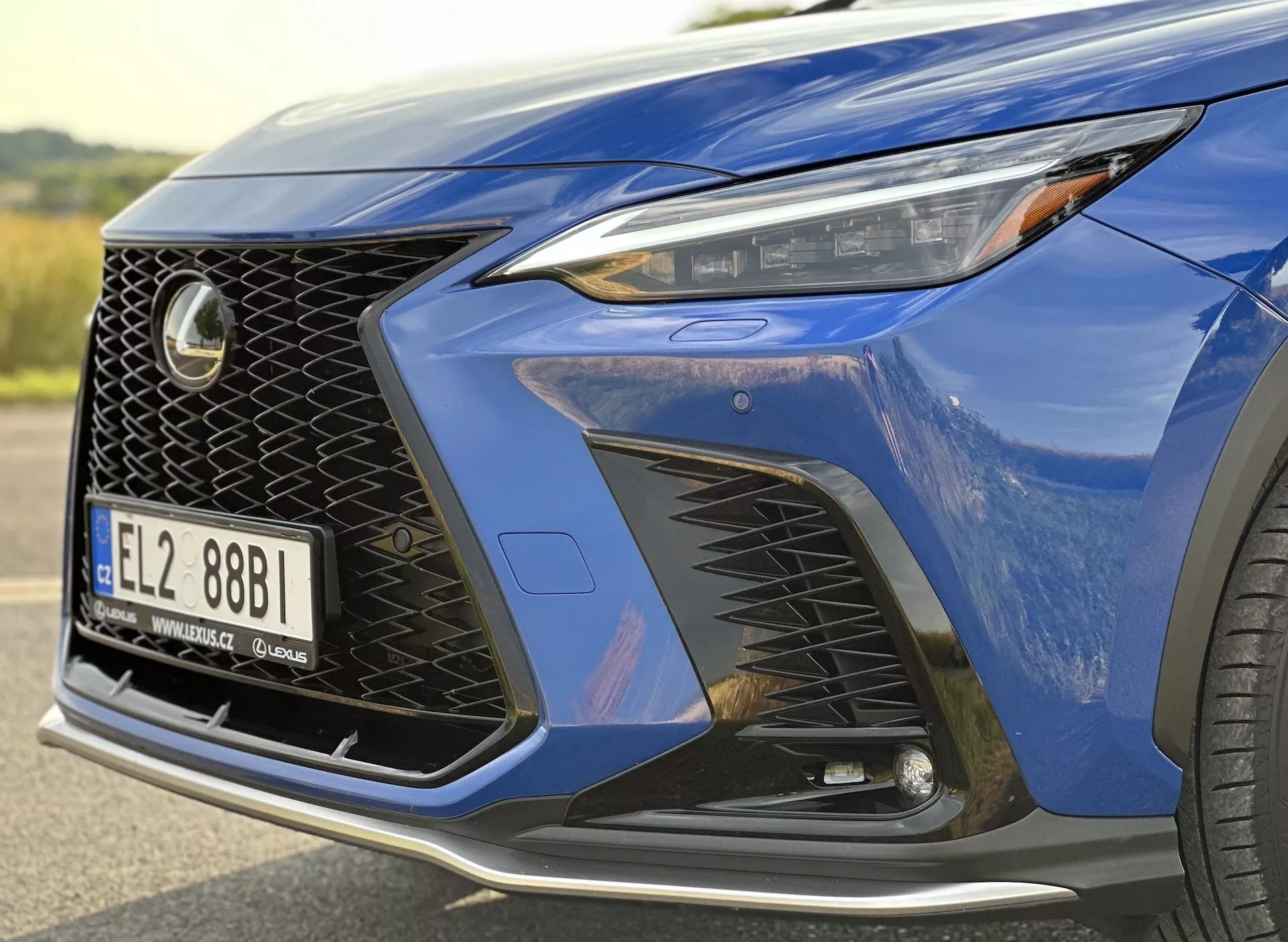 Posnetek sprednjega kota Lexusa NX 2021, ki prikazuje žaromet z LED tehnologijo, značilno mrežasto zasnovo maske in aerodinamični sprednji odbijač z velikimi zračnimi vhodi, poudarjen z globoko modro kovinsko barvo in delčkom luksuznega lita kolesa.
