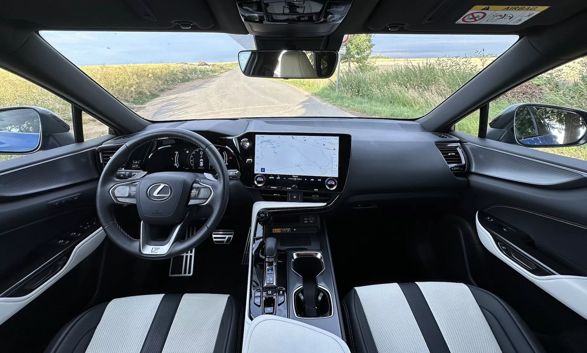 Notranjost Lexusa NX 2021 iz perspektive voznika, ki prikazuje volan na sredini z logotipom Lexus in kontrolnimi gumbi, velik zaslon za infotainment na armaturni plošči, minimalistične kontrole za klimatsko napravo, prestavna ročica avtomatskega menjalnika na sredinski konzoli z držali za pijačo in prostorom za shranjevanje, sedeže oblazinjene z dvobarvnim materialom, in delni pogled na podeželsko pokrajino skozi vetrobransko steklo.