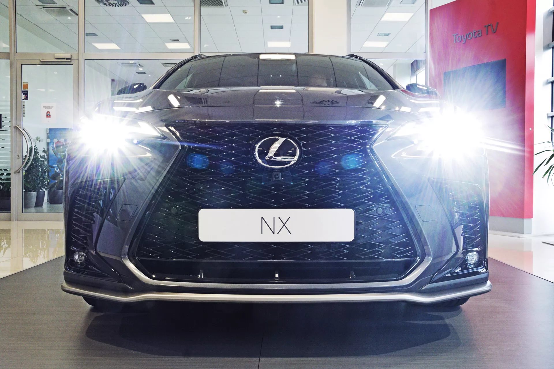 Sprednji pogled na vozilo Lexus NX 2021, fotografirano iz nizkega kota, ki prikazuje njegovo višino in prisotnost na cesti v razstavnem okolju. Slika poudarja značilno vretenasto masko s peščeno uro in temno mrežo, osvetljene kotne LED žaromete z učinki bleščanja, dnevne luči in podrobnosti spodnjega odbijača. Sijoča tla in nosilec registrske tablice z oznako 'NX' prispevajo k luksuznemu in tehnološkemu videzu avtomobila.