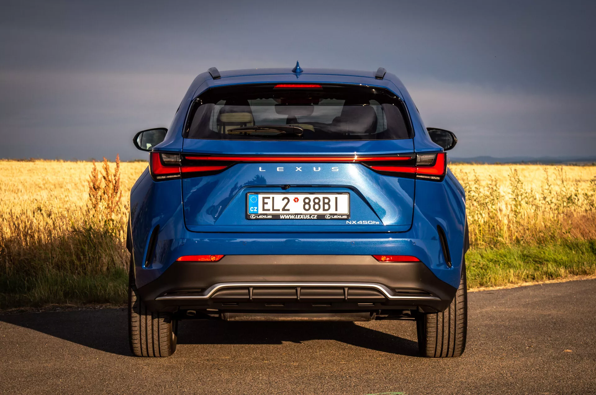 Zadnji pogled na modri SUV Lexus NX 2021 na asfaltni cesti z zlatim poljem v ozadju, ki poudarja hibridno tehnologijo in oblikovne značilnosti