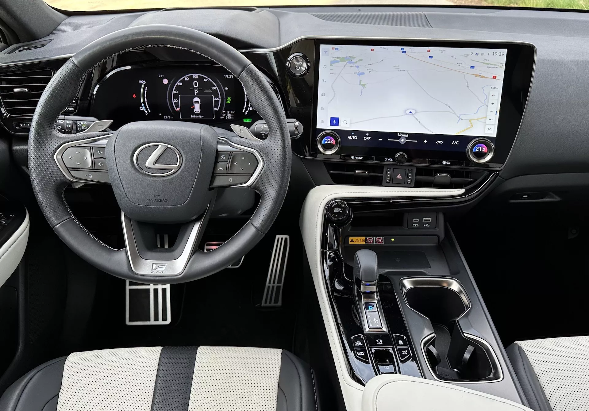 Notranji pogled na Lexus NX 2021, ki prikazuje voznikovo območje in sredinsko konzolo iz dvignjenega kota, z logotipom Lexus na volanu z nameščenimi kontrolniki, osvetljenim digitalnim inštrumentnim sklopom, velikim zaslonom infotainment sistema na dotik z GPS navigacijo, gumbi za nadzor klime in avdio, ergonomskim izbirnikom prestav, kontrolami načina vožnje, luksuznimi materiali na sedežih in dotikalnih točkah, kovinskimi poudarki in vidnimi držali za skodelice in predalom za shranjevanje.