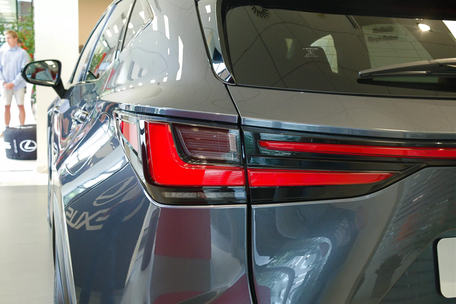Povečan posnetek Lexus NX 2021 iz zadnjega tričetrtinskega kota, ki poudarja eleganten dizajn LED zadnje luči, del zadnjega bočnega panela in del pokrova prtljažnika. Slika prikazuje temno sijočo barvo avtomobila, aerodinamične linije karoserije in namiguje na notranji razstavni prostor z odsevi drugega vozila, arhitekturnih elementov in osebe, ki hodi v ozadju.