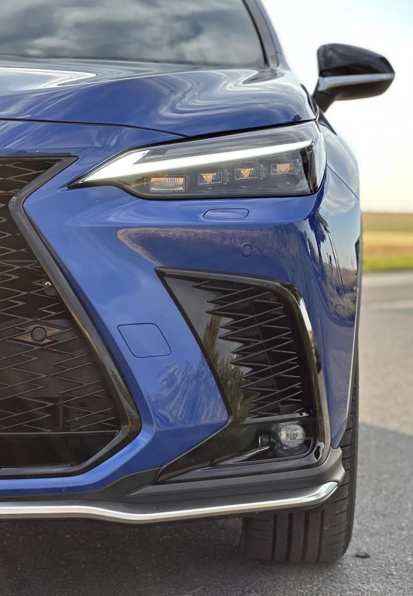 Podroben pogled na sprednji levi kot 2021 Lexus NX v temno modri barvi, ki poudarja napredno zasnovo LED žarometov, zapleten vzorec rešetke, športni sprednji odbijač in možne adaptivne svetlobne in aerodinamske funkcije z bleščečim kovinskim zaključkom