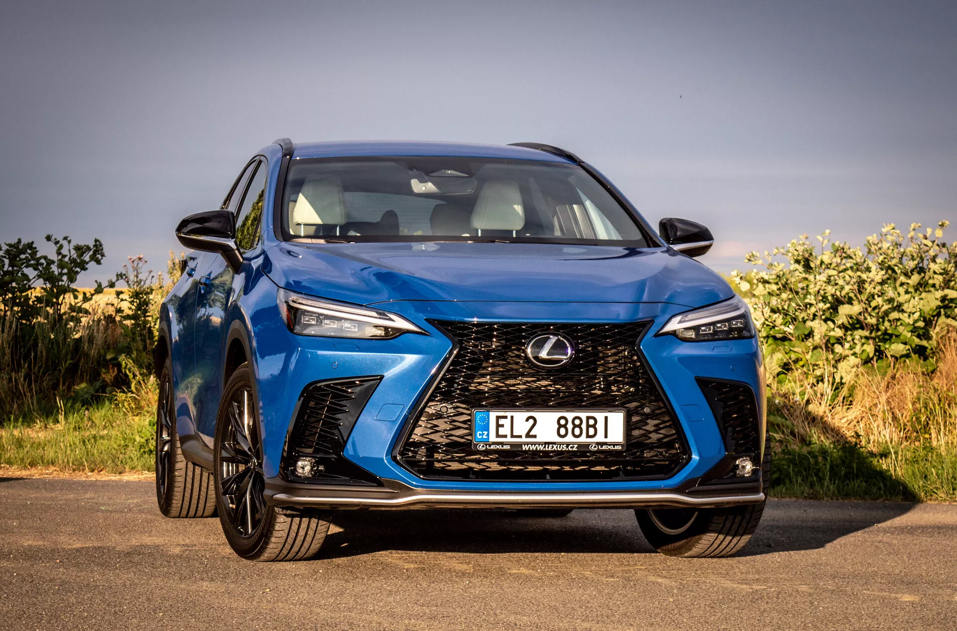 Sprednji kotni pogled na modri SUV Lexus NX 2021, ki poudarja njegov zapleteni vretenasti žaromet, kotne LED žaromete, sijoč zaključek in aerodinamično zasnovo, parkiran na prostem pod jasnim nebom
