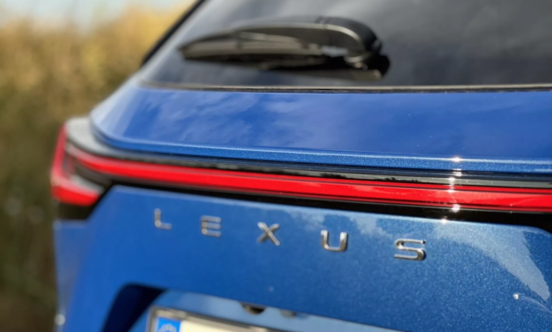 Pogled od blizu na zadnji del znamenja vozila Lexus NX 2021 z srebrnim znakom Lexus, rdečo svetlobno črto nad imenom na metalik modrem telesu pri dnevnih svetlobnih pogojih