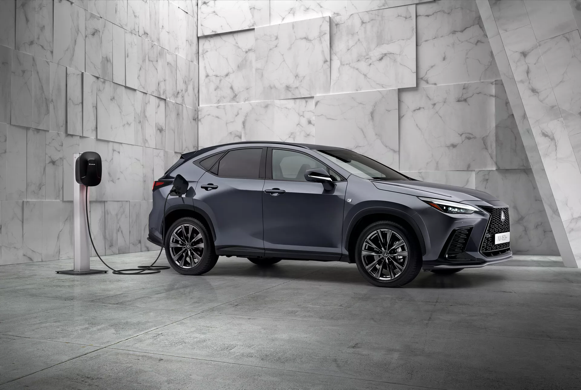Ta slika prikazuje Lexus NX 2021, sodoben kompaktni SUV z elegantnim in športnim dizajnom, fotografiran iz treh četrtin spredaj. Vidni sta leva stran in deloma tudi sprednji del vozila, nameščena diagonalno v minimalističnem okolju, okrašena z velikimi svetlimi marmoriranimi ploščicami, kar nakazuje na premijsko vzdušje. Lexus NX se ponaša z značilnimi kotnimi LED žarometi in izrazito sprednjo mrežo, kar poudarja njegovo sofisticiranost. Vidna so aerodinamično oblikovana bočna ogledala, zapleteni večkraki aluminijasti platišči in sploščeni ročaji vrat, kar poudarja športni in napredni dizajn. Pomembna podrobnost je povezava avtomobila z električno polnilno postajo, kar nakazuje, da gre za električno vozilo ali priključni hibrid. Prednji blatnik vključuje polnilno odprtino, kar označuje električne zmožnosti vozila. Ta statična slika se osredotoča na prikaz dizajna vozila in tehnoloških funkcij v uravnoteženi in mirni postavitvi z obilico negativnega prostora, kar ponazarja ekskluzivnost in inovativnost v avtomobilskem dizajnu.
