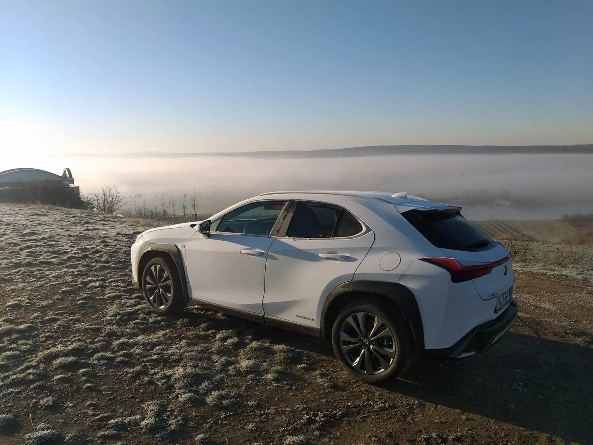 Crossover SUV Lexus UX 2019 parkiran na travniku pokritem z zmrzaljo z ozadjem meglenega pokrajinskega, prikazuje profil vozila iz zadnjega tristranskega kota, poudarjajoč stranske značilnosti linij, obokane kolotekov, zožujočo se strešno linijo in lita platišča.