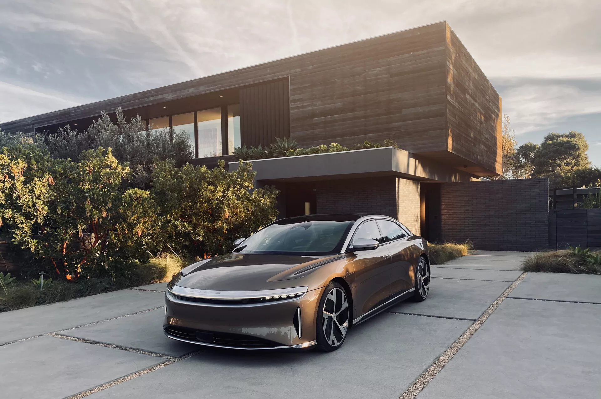 Slika 2022 Lucid Air sodobnega sedana, parkiranega pred sodobno hišo. Posneta iz nizkega, sprednjega trehčetrtinskega zornega kota, ki prikazuje njegov eleganten, aerodinamičen dizajn z podaljšanim pokrovom motorja in subtilno mrežico. Glavni poudarki vključujejo tanke LED luči, velika izrazita kolesa in odsotnost stranskih ogledal, kar nakazuje na uporabo kamero kot sistem za vzvratni pogled. Kovinska temna barva in izpopolnjeno okolje poudarjata njegov luksuzen občutek, kar nakazuje, da je to električno vozilo namenjeno premožnemu tržnemu segmentu.