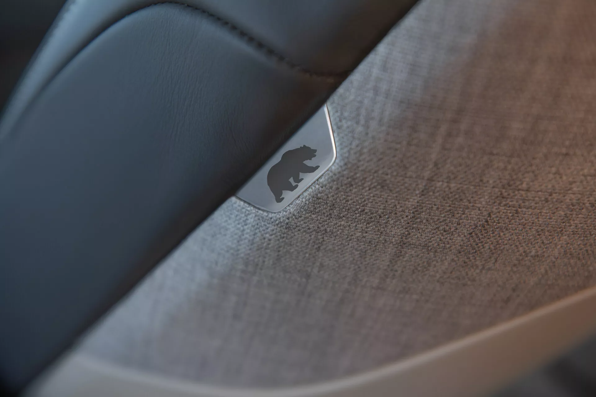 Povečava, selektivno izostrena slika notranjosti vozila Lucid Air 2022 (1. generacija), ki poudarja podrobnosti vrat ali armaturne plošče. Kovinski emblem ali značko, ki odbija svetlobo, je vidno postavljen na teksturirano blago ali mehko plastiko z detajli šivov. Gladka, temnejša snov, verjetno črno usnje, kontrastira s teksturiranimi elementi, kar prikazuje kakovostno izdelavo in visokokakovostno notranjost avtomobila.