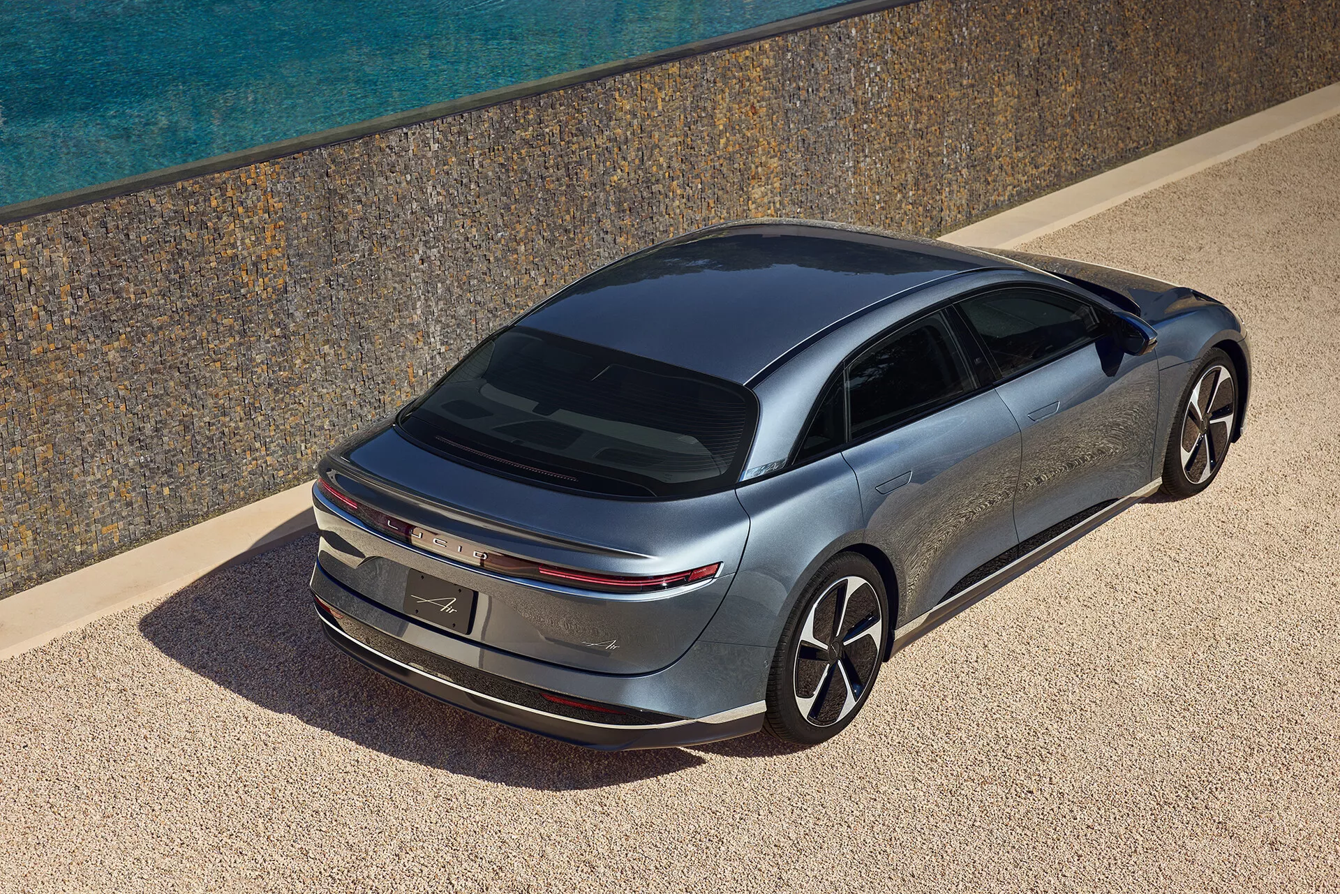 Visoko zorni posnetek elegantnega sedana Lucid Air 2022, gledanega od zgoraj iz zadnje četrtine. Vozilo ima silhueto podobno kupeju z nagnjeno strešno linijo, tankimi horizontalnimi zadnjimi lučmi in kovinskim zaključkom barve. Okolje vključuje morje in luksuzno rezidenco, kar dodaja občutek vrhunske kakovosti. Minimalistična zunanja zasnova avtomobila, velika večkraka platišča in zatemnjena okna nakazujejo osredotočenost na aerodinamiko in sodobno tehnologijo, kar lahko namiguje na električni pogon.