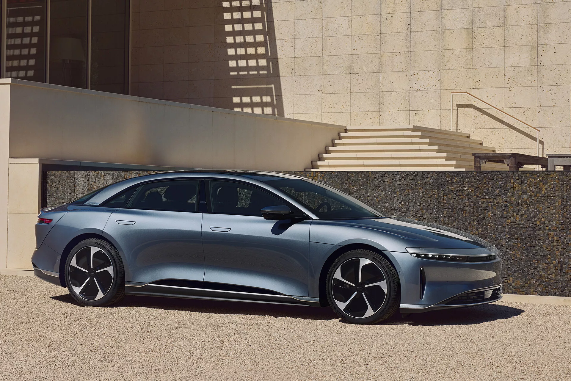 Stranski profil Lucid Air 2022, sodobnega elegantnega sedana, posnetega na prostem v urbanem okolju z arhitekturnimi elementi, ki mečejo sence. Leva stran avtomobila prikazuje njegov eleganten, aerodinamični dizajn z nizko strešno linijo, vgradnimi ročaji vrat, velikimi večkrakimi kolesi in tankimi bočnimi ogledali. Slika poudarja dolžino avtomobila, od sprednjega do zadnjega odbijača, s prednjim A-stebrom, ki daje vtis prostorne notranjosti, in dinamično karakterno linijo na karoseriji.