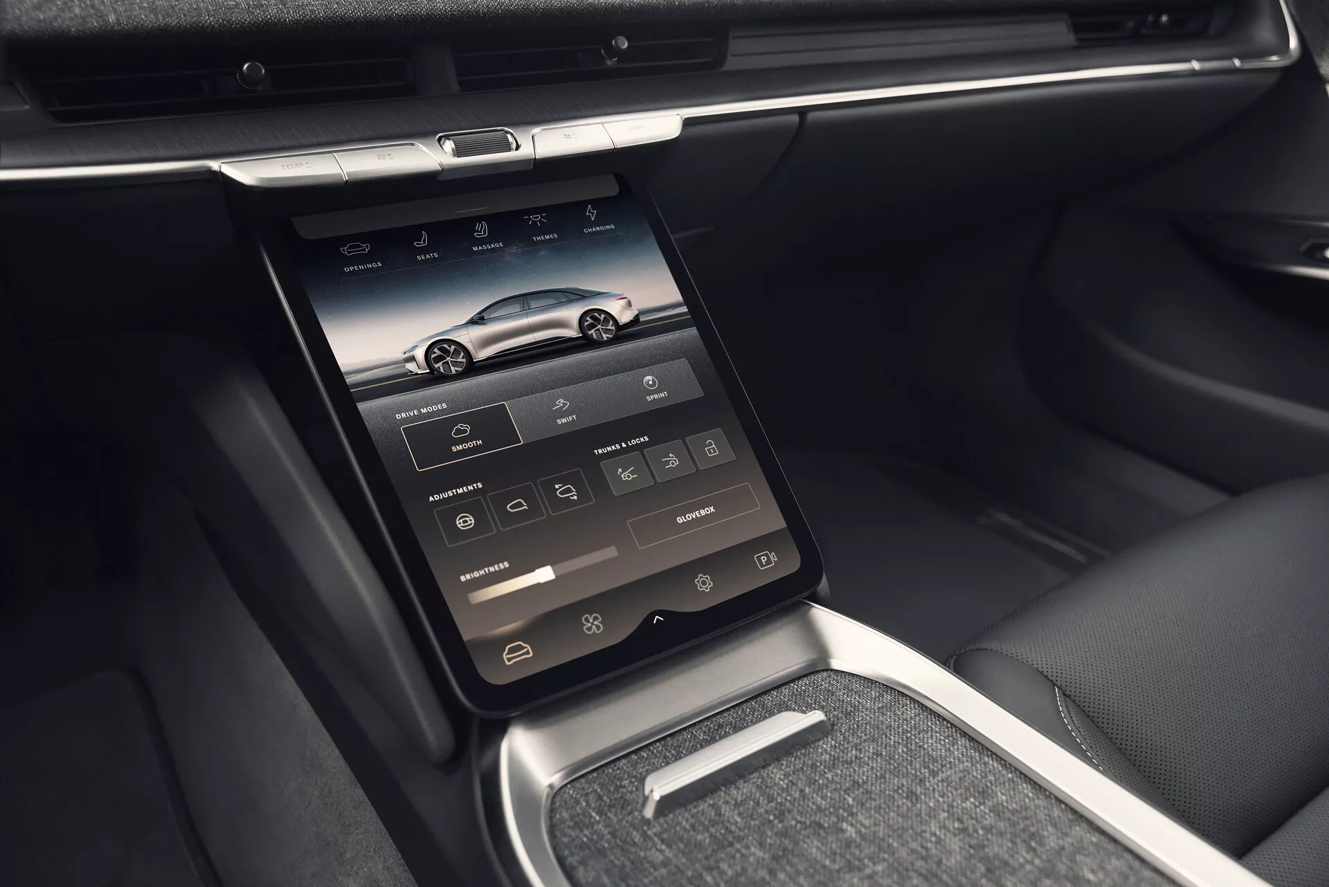 Pogled v notranjost modela Lucid Air 2022 (prva generacija) prikazuje sredinsko konzolo z vgrajenim pokončno nameščenim zaslonom na dotik, na katerem so ikone za navigacijo, telefon, medije, nadzor klime in nastavitve vozila. Fotografija, posneta iz rahlo levo nagnjene perspektive, poudarja napredne tehnološke funkcije z zračniki nad zaslonom, sijočimi kromiranimi ali kovinskimi okrasnimi elementi, elegantno armaturno ploščo in mešanico usnjenih ter kovinskih površin. Slika tudi podrobno prikazuje manjšo polico pod zaslonom na dotik, kovinsko prestavno ročico in usnjene rokonaslone ter stranice sedežev, poudarja luksuzno in udobno notranjost.