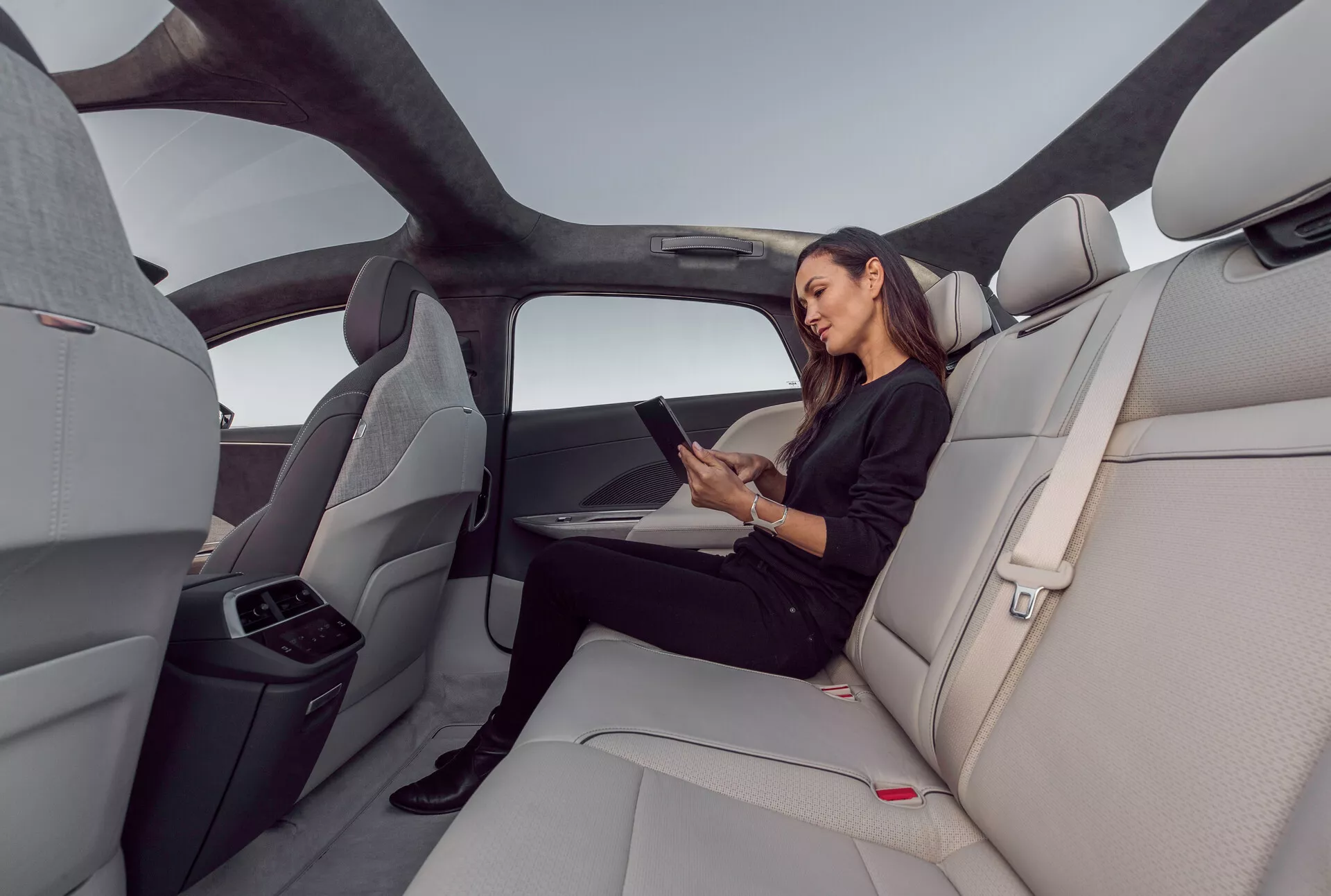 Notranji pogled zadnjega sedežnega prostora vozila Lucid Air 2022, ki poudarja prostorno, luksuzno kabino s svetlo sivimi, oblikovanimi usnjenimi sedeži. Slika zajema širokokotni posnetek z desne strani, ki prikazuje panoramsko streho, ki se razteza čez zadnji prostor, ergonomsko oblikovane sedeže in zložljiv osrednji naslon za roke z držali za pijačo. Ženska sedi udobno in uporablja tablico, kar kaže na možne povezovalne funkcije. Elegantna, minimalistična notranjost z rdečimi gumbi za sprostitev varnostnega pasu in vodoravnimi črtami na armaturni plošči in vratnih panelih, ki povečujejo občutek širine in odprtosti.