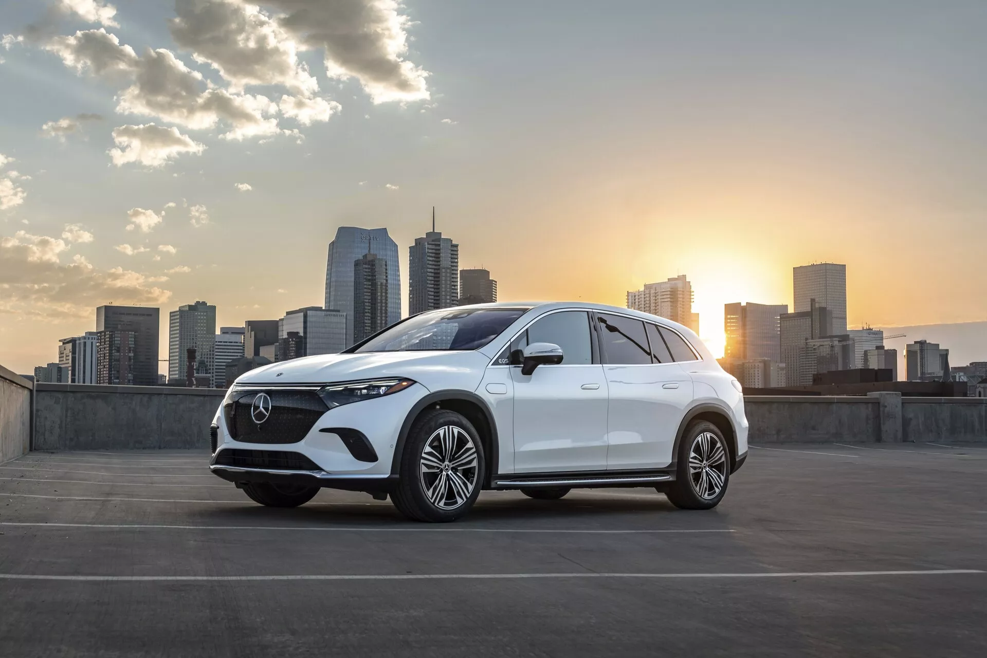 Rafiniran bel Mercedes EQS-SUV 2022 parkiran s panoramo mesta in zahajajočim soncem v ozadju. Kot sprednjega tričetrtinskega pogleda prikazuje vozilo z značilno izrazito masko z natančnim mrežastim vzorcem in centralnim emblemom, elegantnimi LED žarometi, dvojnimi zračniki ali prezračevalnimi odprtinami in velikimi večkrakimi zlitinami platišč. Nežna padajoča linija strehe in povečana višina od tal nakazujeta obliko SUV. Bočna ogledala verjetno vsebujejo tehnološke funkcije, kot so integrirani smerniki ali kamere za spremljanje mrtvega kota. Slika je lepo osvetljena z mehkimi poudarki zahajajočega sonca, s poudarkom na oblikovnih linijah vozila. Urbano okolje z nebotičniki dodaja sodoben in prefinjen pridih, nakazuje ciljno skupino mestnih potrošnikov, ki cenijo slog in napredno tehnologijo.