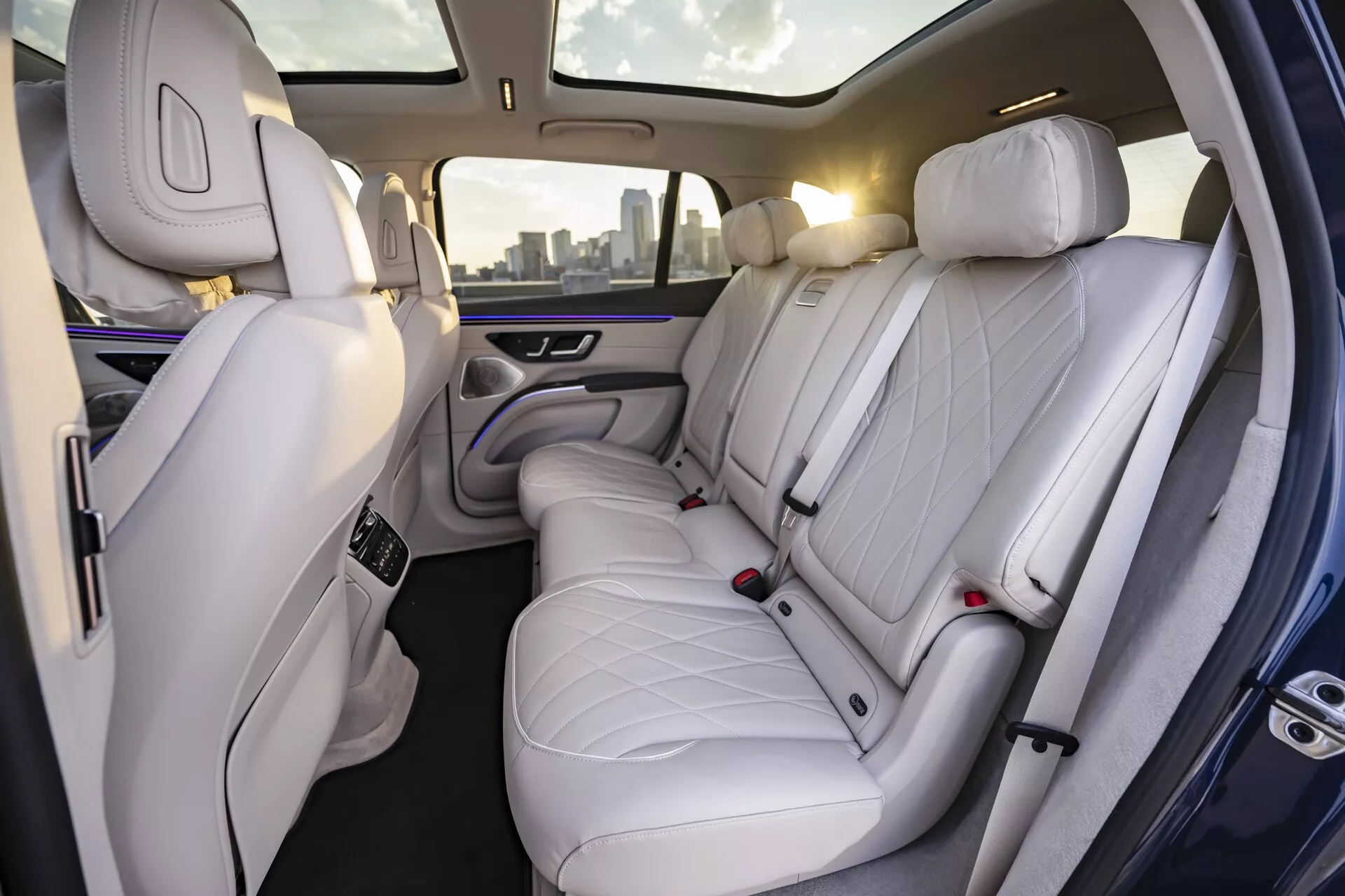 Pogled na notranjost Mercedes EQS-SUV 2022, ki prikazuje luksuzni zadnji potniški predel. Posnetek, narejen pod kotom, rahlo nagnjenim v desno, razkriva visokokakovostne bele ali svetlo sive usnjene sedeže z oblazinjenim vzorcem diamanta. S tremi vzglavniki sedeži nudijo dovolj prostora, kar kaže na poudarek na udobju in eleganci. Vidna je tudi zadnja stran sedeža sopotnika, prav tako prevlečena s premium usnjem. Sofisticirana ambientalna osvetlitev z nežnim vijoličnim sijajem vzdolž vratnih plošč in centralnega naslona za roke izboljšuje občutek premije. Centralni naslon za roke vključuje kontrolne enote, verjetno za nastavitve klime in sedežev, ter vsebuje dva držala za pijačo.