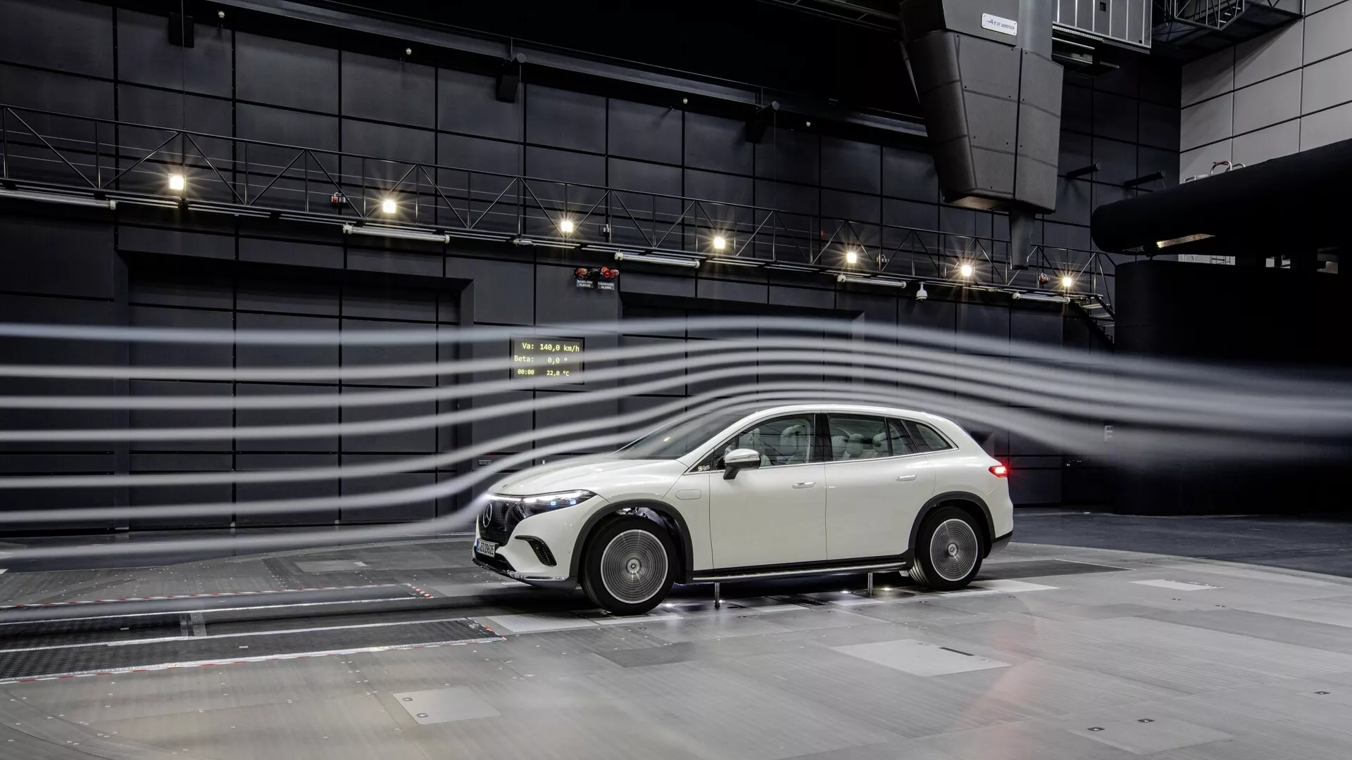Slika prikazuje stranski profil vozila Mercedes EQS-SUV 2022, ki poudarja njegovo moderno zasnovo v nadzorovanem testnem okolju. Model kombilimuzine ali karavana je poudarjen z značilno sprednjo režo, elegantnimi stranskimi ogledali, večkrakimi lita platišči, integriranimi meglenkami v sprednjem odbijaču in anteno nameščeno na strehi. Aerodinamične linije in vizualizirani tokovi zraka okoli avtomobila nakazujejo poudarek na učinkovitosti porabe goriva in zmogljivosti, pri čemer mokra površina, ki se odseva avtomobil, verjetno nakazuje testiranje interakcije s površino ceste. Konfiguracija nakazuje napredno tehnologijo in aerodinamične teste, potencialno za funkcije, kot so aktivna aerodinamika, sistemi za pomoč vozniku in nadzor vleke.