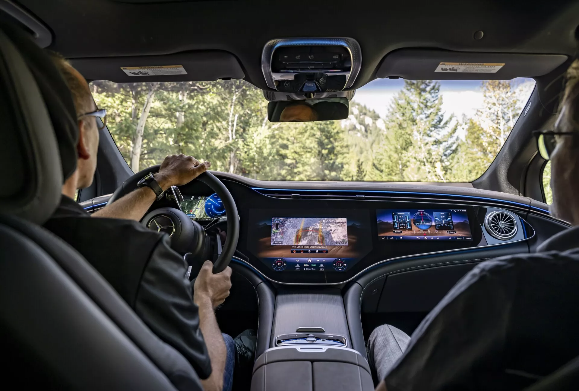 Pogled v notranjost avtomobila Mercedes EQS-SUV 2022 s perspektive srednjega zadnjega sedeža, ki prikazuje sodobno armaturno ploščo z neprekinjenim digitalnim zaslonom, sodoben volan z vgrajenimi kontrolami, razkošne svetle usnjene sedeže in dvignjeno sredinsko konzolo. Vetoransko steklo ponuja pogled na cesto obdano z gozdom, kar nakazuje, da je avtomobil v gibanju.