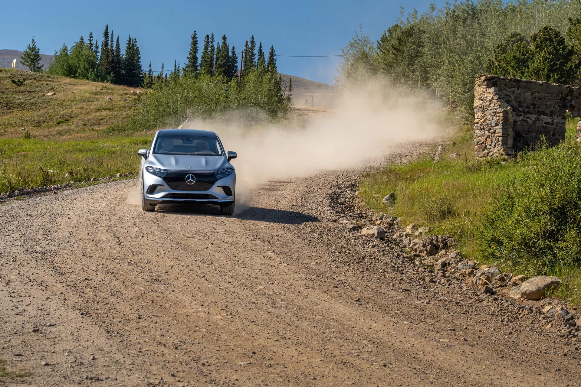 Mercedes EQS SUV 2022 – Sodobni hatchback ali kompaktni SUV, ki vozi proti kameri po makadamski cesti, za seboj pušča oblak prahu. Avto je postavljen v sredino in jasno prikazuje sprednjo masko, žaromete, pokrov motorja, vetrobransko steklo in del strehe. Okolje nakazuje na podeželsko ali terensko okolje s travo na obeh straneh, jasno nebo in ostanke stare kamnite strukture v ozadju.