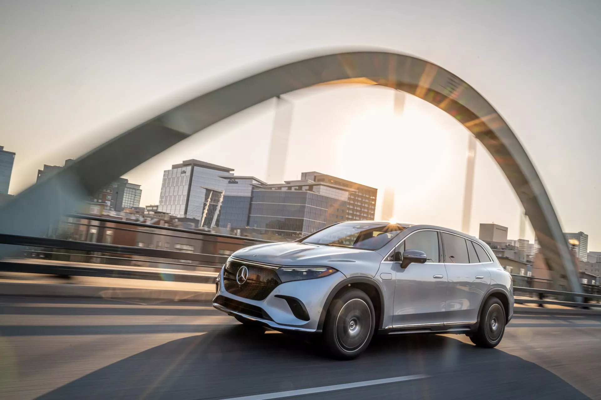 Slika prikazuje srebrni Mercedes EQS-SUV 2022, ki vozi po dvignjenem mostu. Posneta iz nizkega kota, tričetrtinski sprednji pogled poudarja sodobno obliko SUV. Voznikova stran razkriva izrazito kromirano masko z emblemom, elegantne linije, stilizirane LED žaromete in zameglitev gibanja na kolesih, kar pomeni hitrost. Sijoča sončna svetloba in nejasna silhueta mesta dodajata dramatičen učinek, kar poudarja razkošje in napredno inženirstvo.