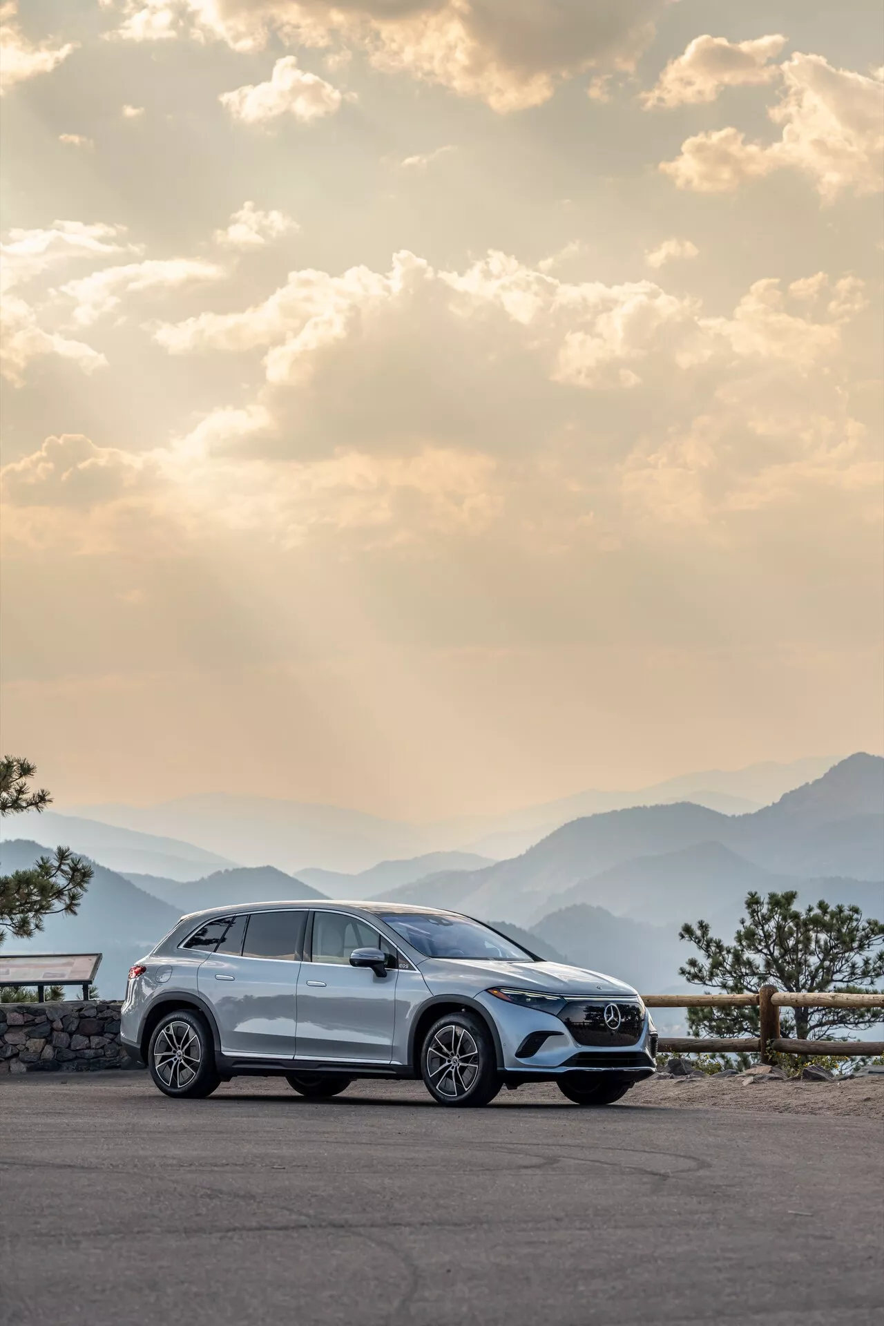 Moderen Mercedes EQS-SUV 2022 parkiran na slikovitem razgledišču, poudarja njegov desni stranski profil z aluminijastimi platišči, kljukami vrat, stranskimi ogledali, okenskimi obrobami in kolesi, rahlo zavrtimi v levo. Posneto ob zori ali mraku s toplo, razpršeno svetlobo, s panoramskimi gorskimi verigami v ozadju in rahlo oblačnim nebom, poudarja luksuz, pustolovščino in visoko kakovostno obrt.