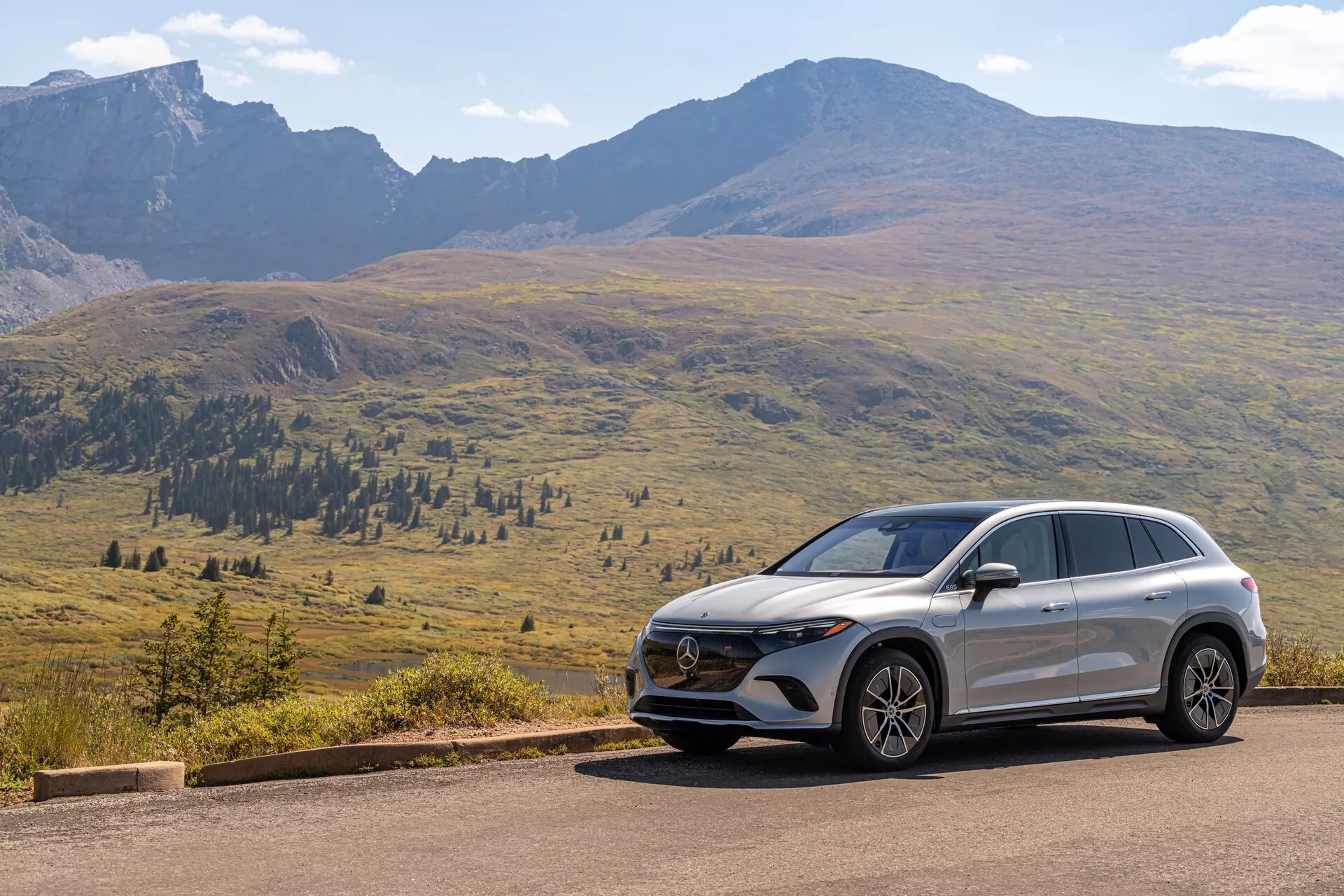 Slika prikazuje sodoben SUV Mercedes EQS-SUV 2022, postavljen na cestno asfaltno pot z slikovitim gorskim ozadjem pod jasnim nebom z razpršenimi oblaki. Pogled iz treh četrtin spredaj razkriva ključne elemente, kot so maska z emblemom, napredna skupina žarometov in oblikovanje prednjega odbijača z senzorji ali kamerami. Aerodinamične linije na pokrovu motorja in ob straneh, skupaj s stranskim profilom katerega so vidna lita platišča, stranska ogledala, značajske linije ter naklon strehe, poudarjajo oblikovalske in tehnične značilnosti vozila.
