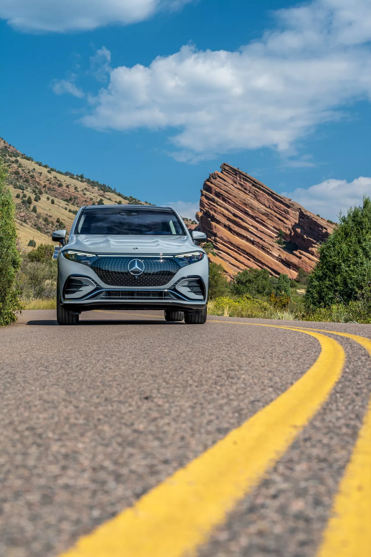 Nizkokotni posnetek Mercedesa EQS SUV 2022, ki se približuje na ovinkasti cesti, prikazuje drzno sprednjo masko z velikim emblemom zvezde, zapleten dizajn v obliki diamanta, LED žaromete in dnevne luči, v ozadju jasno modro nebo in razgiban naravni teren s slojevitimi skalnimi formacijami.